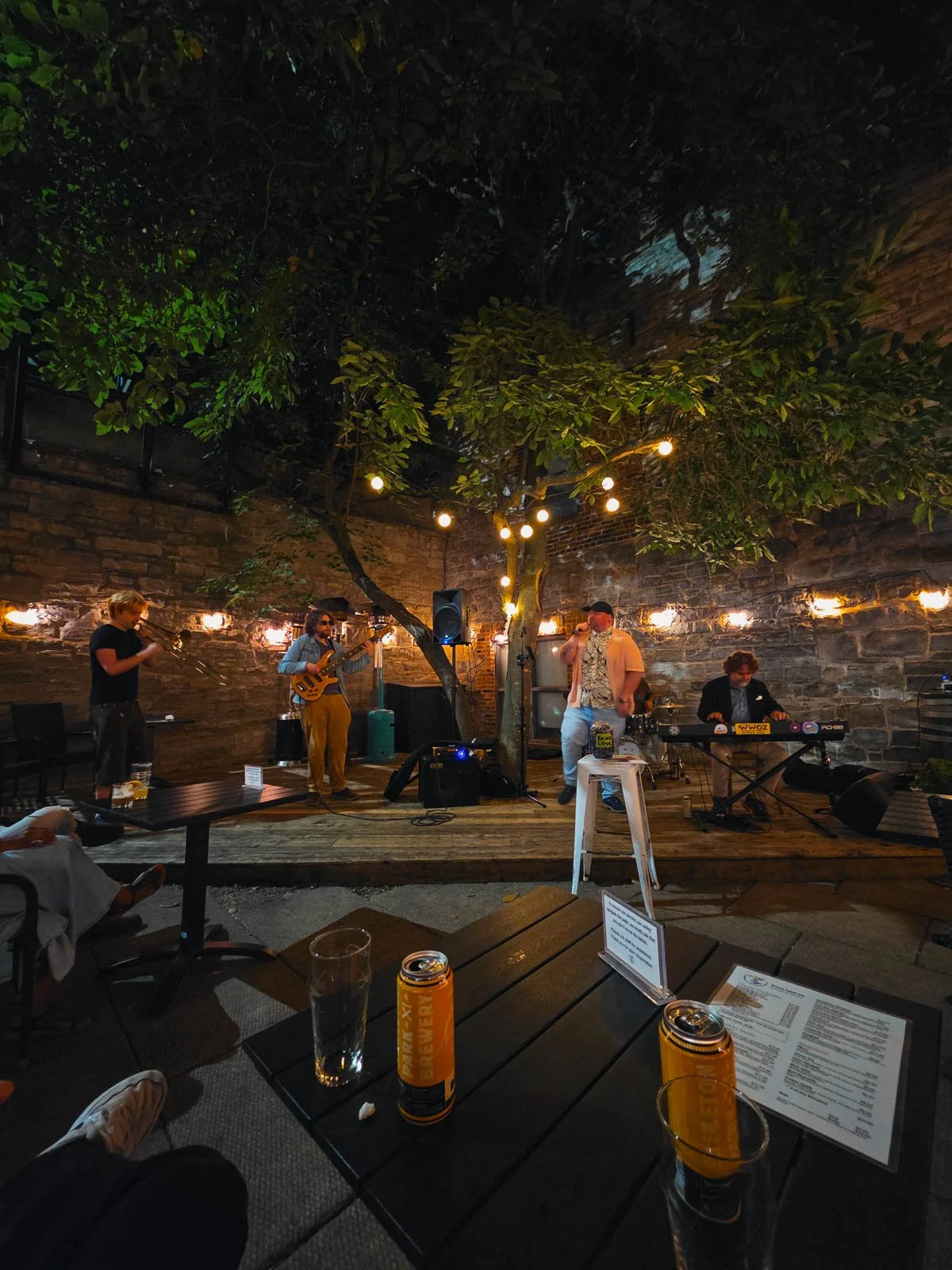 A small outdoor stage in the courtyard of a bar with a live band playing at night with a grey stone walled background