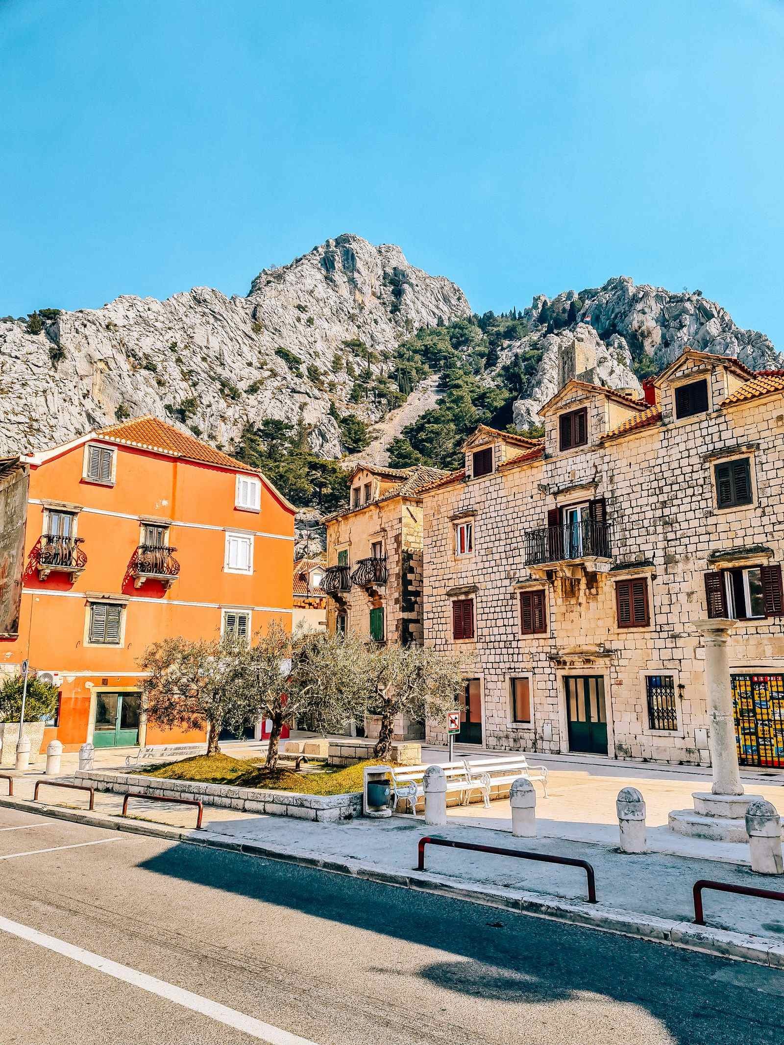 A small square area in the town of Omis, the building on the left is orange, the one on the right is stone and a stone mountain towers in the background with blue sky