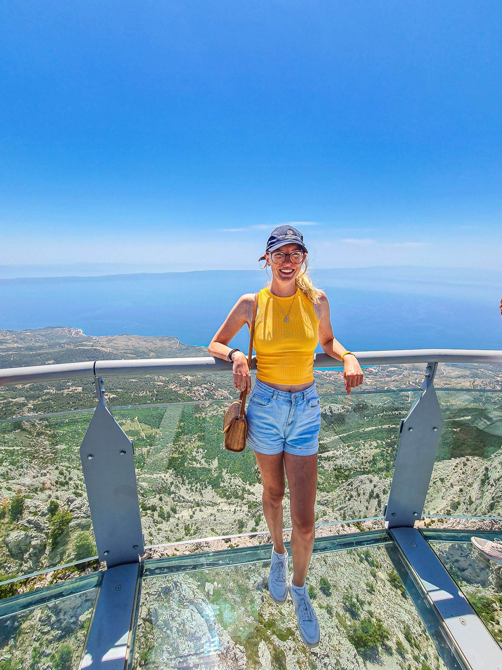 girl in orange top and denim shorts standing on a glass bridge above cliff drop and view of the coast in the distance
