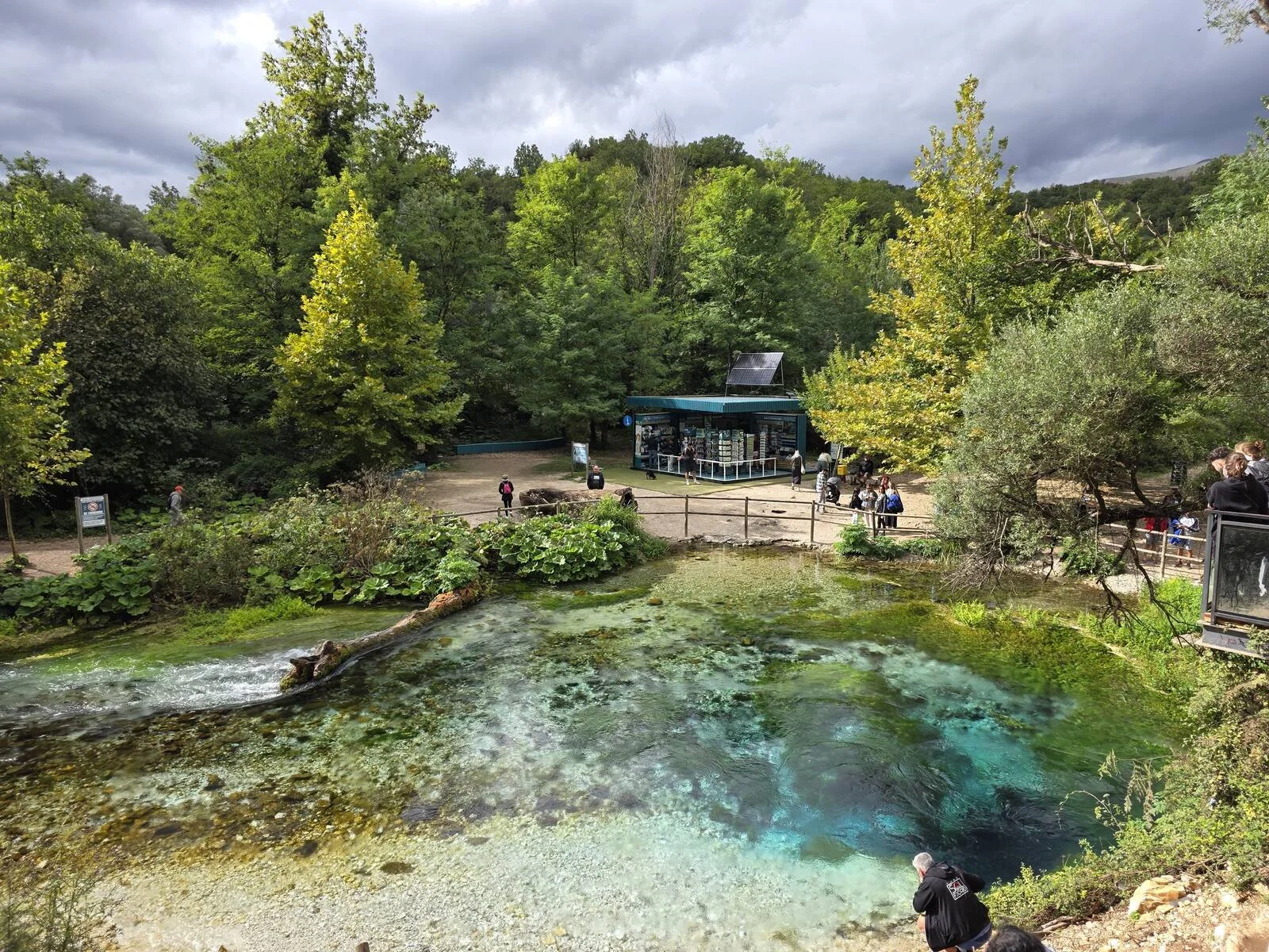 a natural water spring with vibrant green and blue colours surrounded by green trees and a pedestirian walkway on the far side