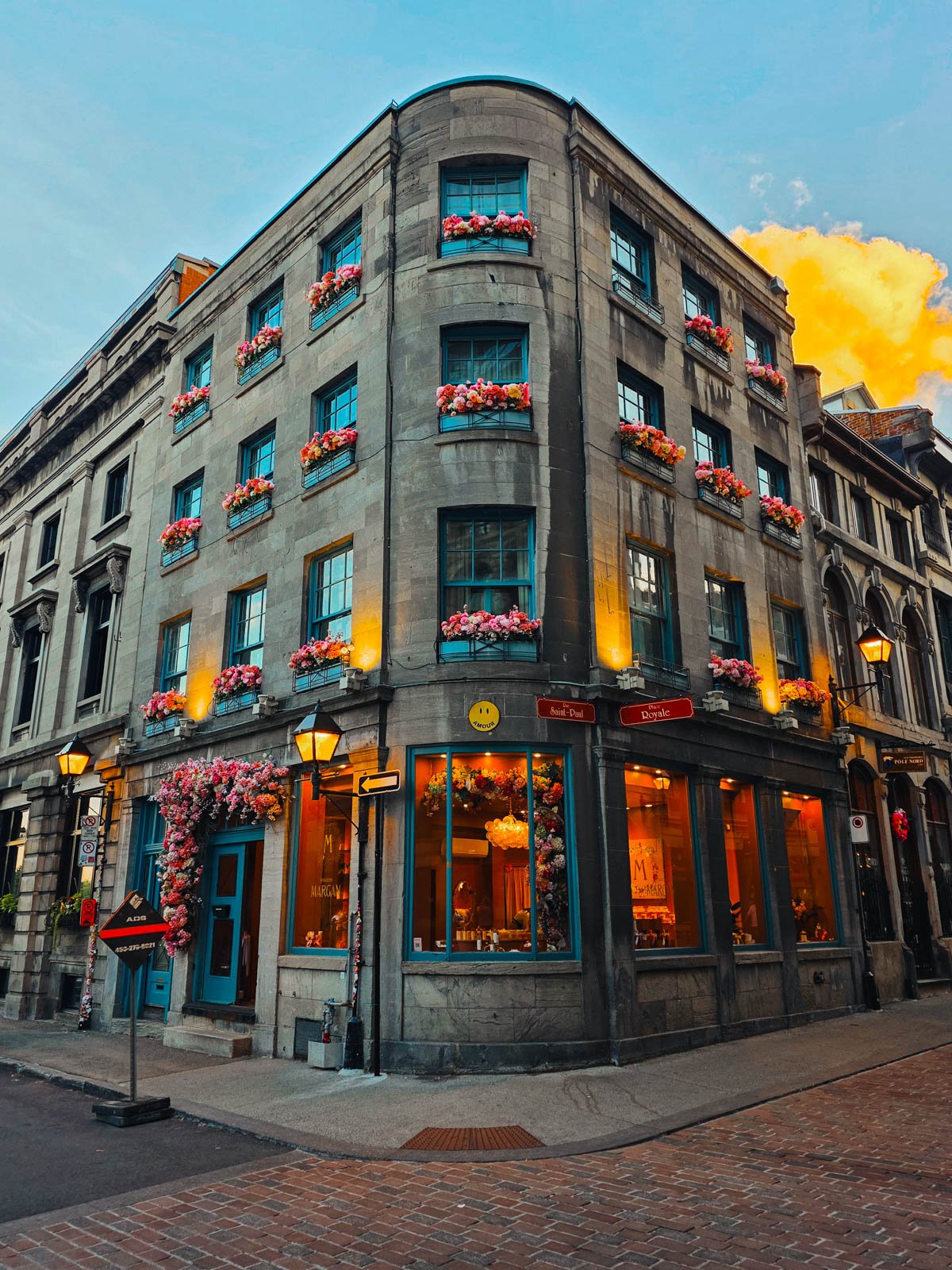 A curved corner building made of stone and with pink flowers in every window box. The interior is warmly lit, this is a building in Old Montréal