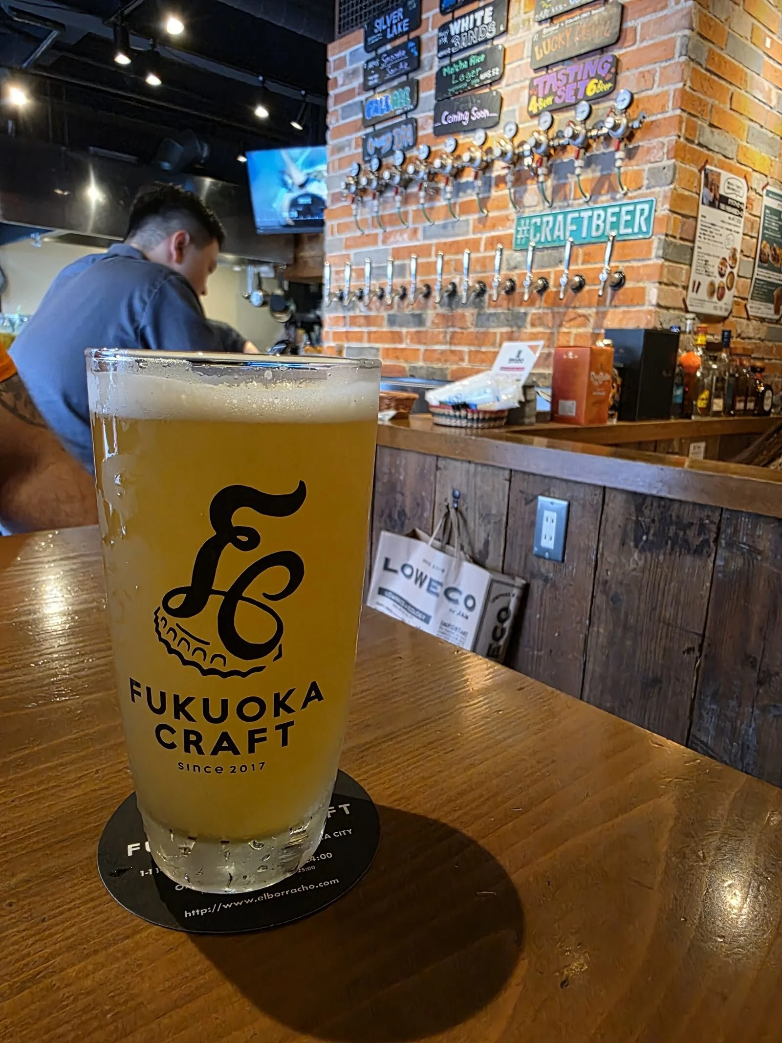 a pint of beer that has "fukuoka craft" written on the glass and many beer taps on the brick wall bar behind