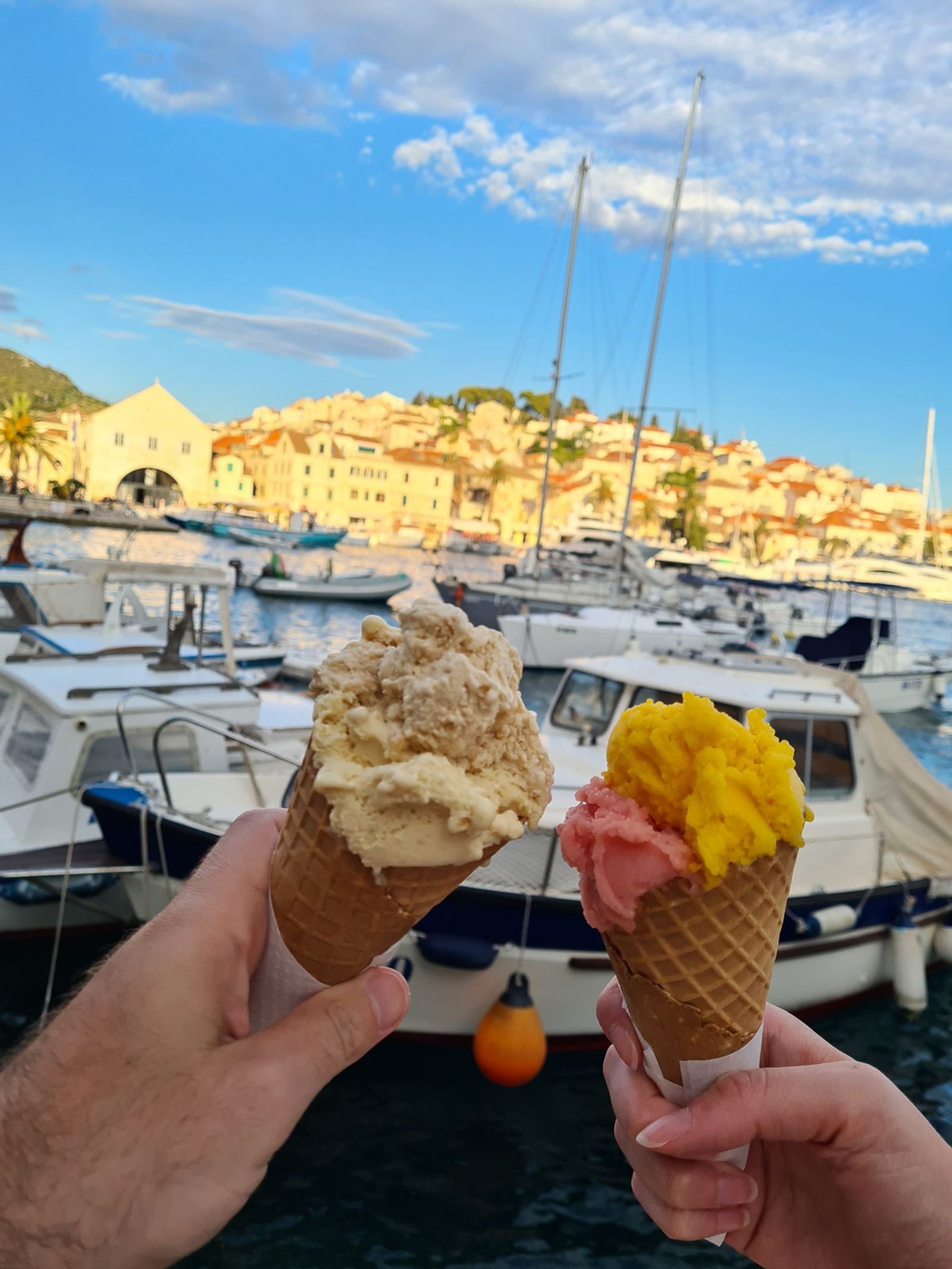 Two hands holding cones of ice cream, one is vanilla, one is pink and yellow. A blurred town in the background
