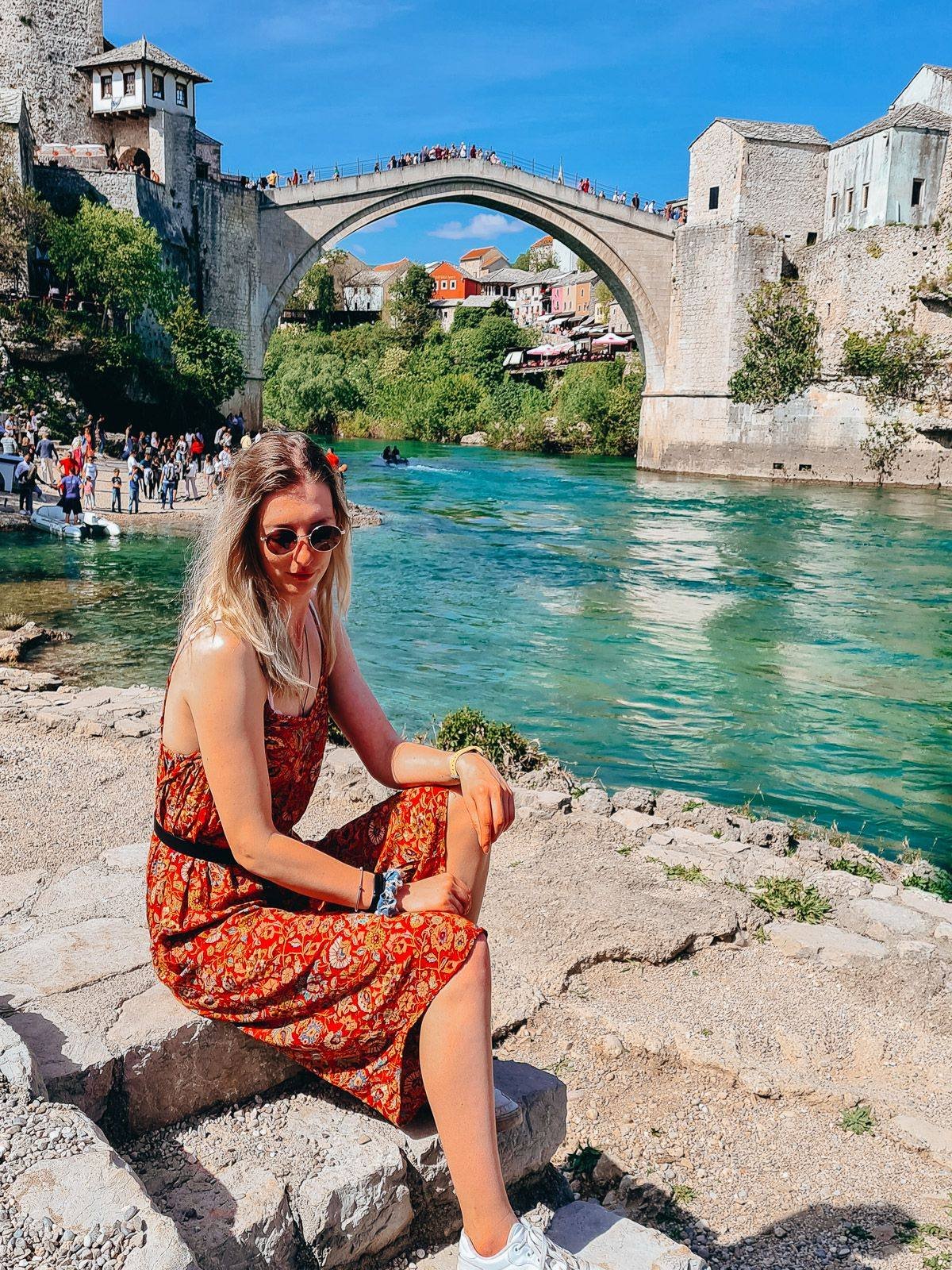 girl sitting on a rock by a river with the famous domed bridge of Mostar in the background