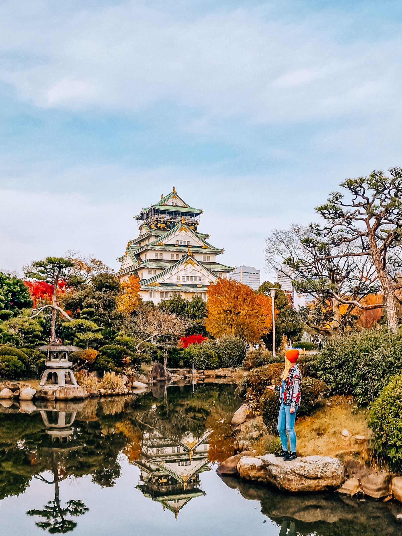 Helena standing on the rock on the edge of a pond in a Japanese garden, a Japanese castle is seen towering above the trees with white walls and green and gold roof.  It is reflected in the pond along with the autumn foliage in the garden