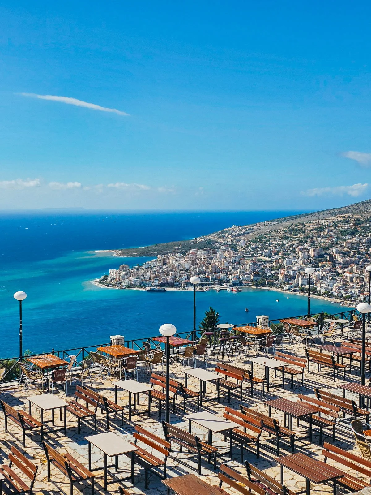 A outdoor seating area with many table a chairs with a view of a seaside town and clear blue water