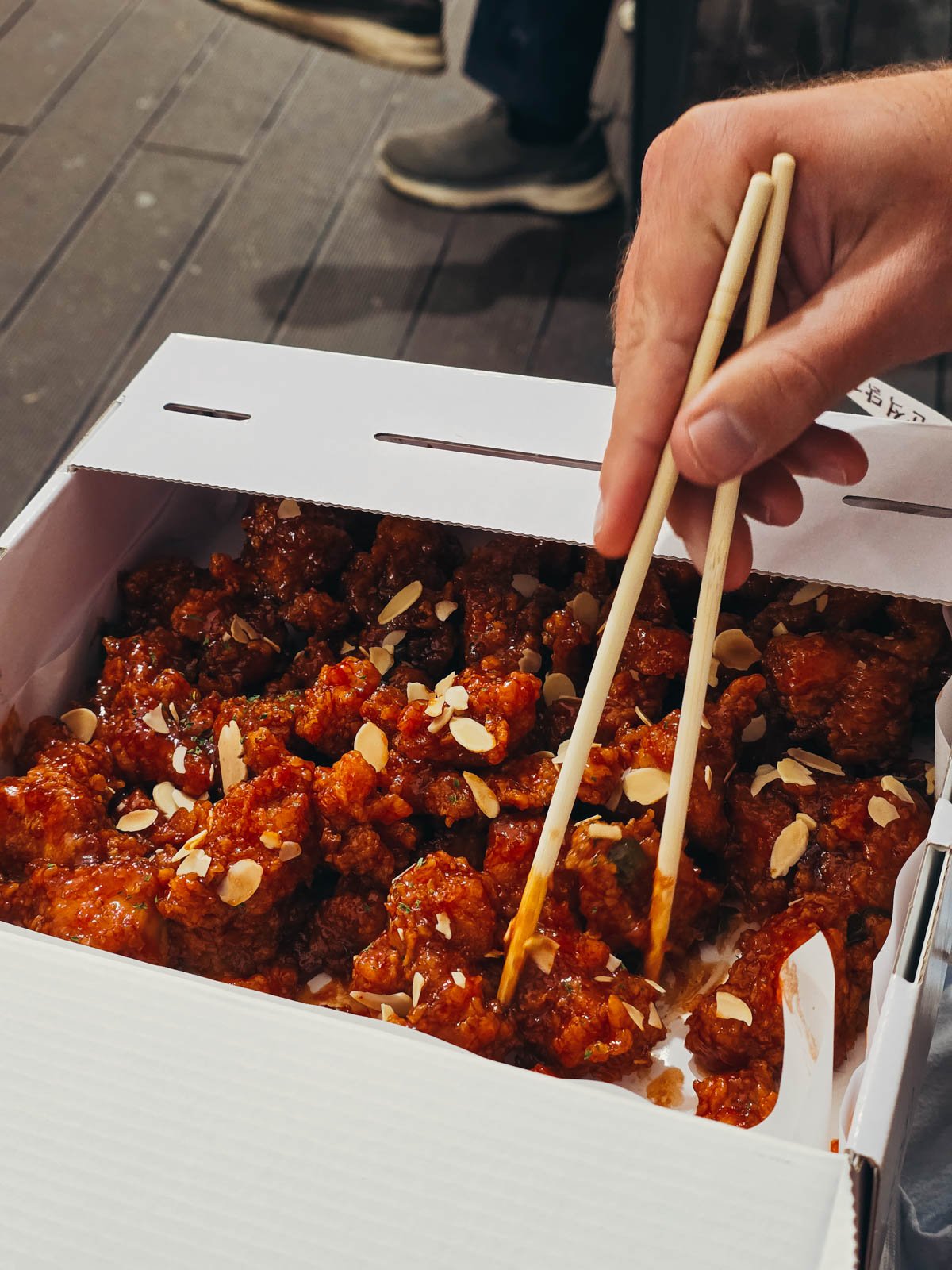A white cardboard box full of sticky fried chicken and two chopsticks about to pick up a piece