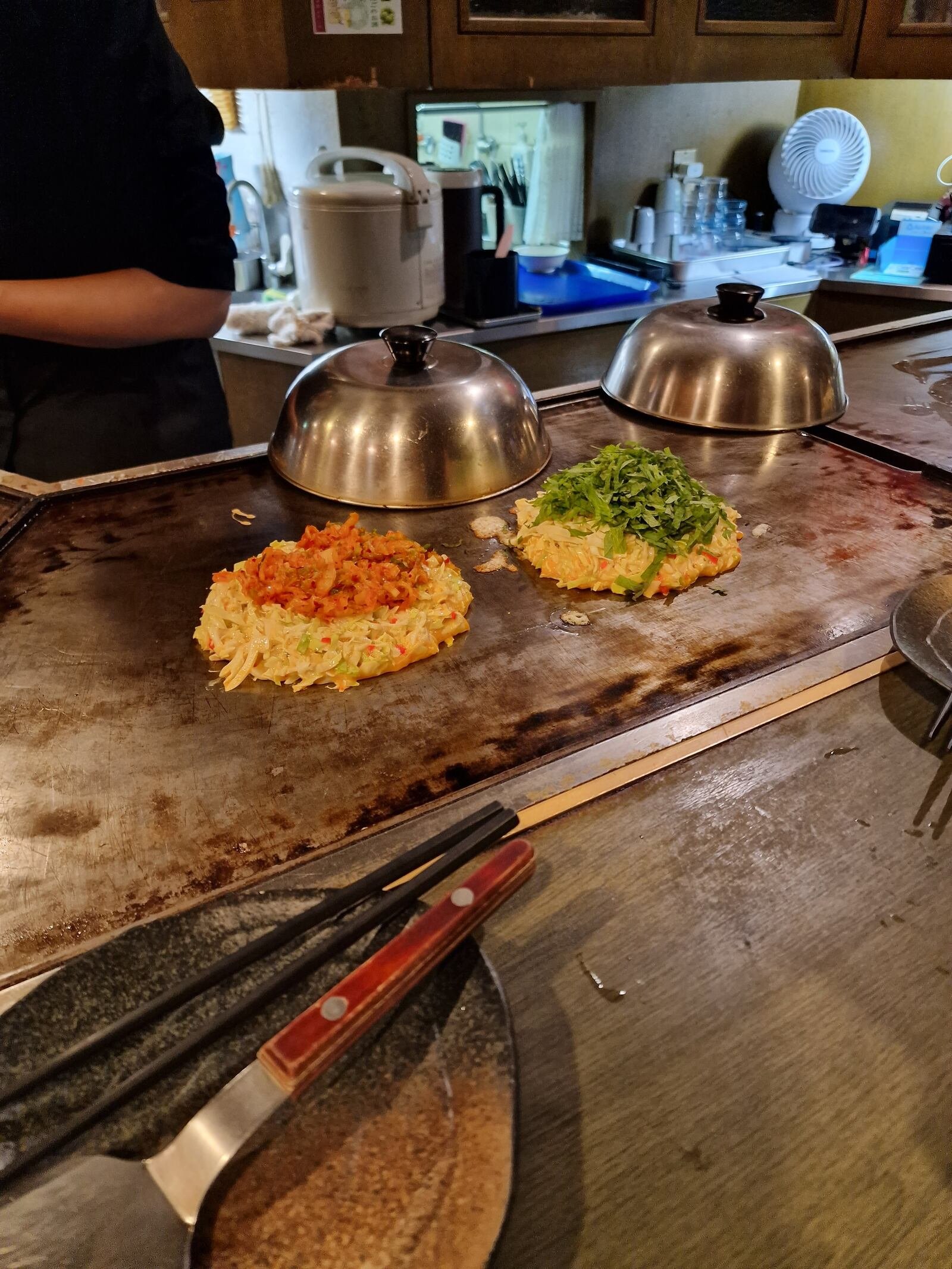 two savory pancakes one with a red topping and one with a green topping cooking on a hot plate in a restaurant