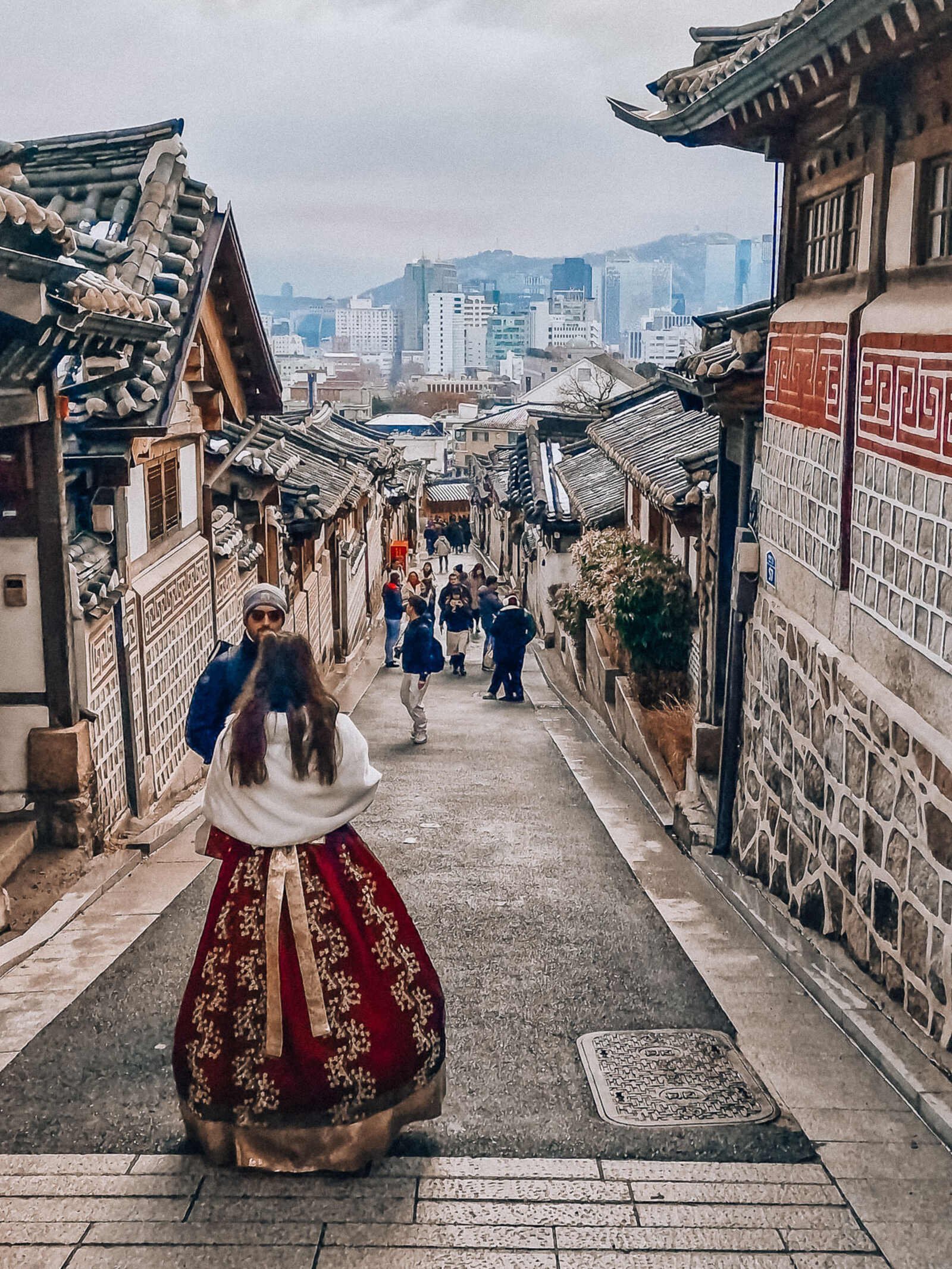 a woman in a traditional red hanbok korean dress looking down a street on a hill in a traditional village. Seoul city is visible in the distance