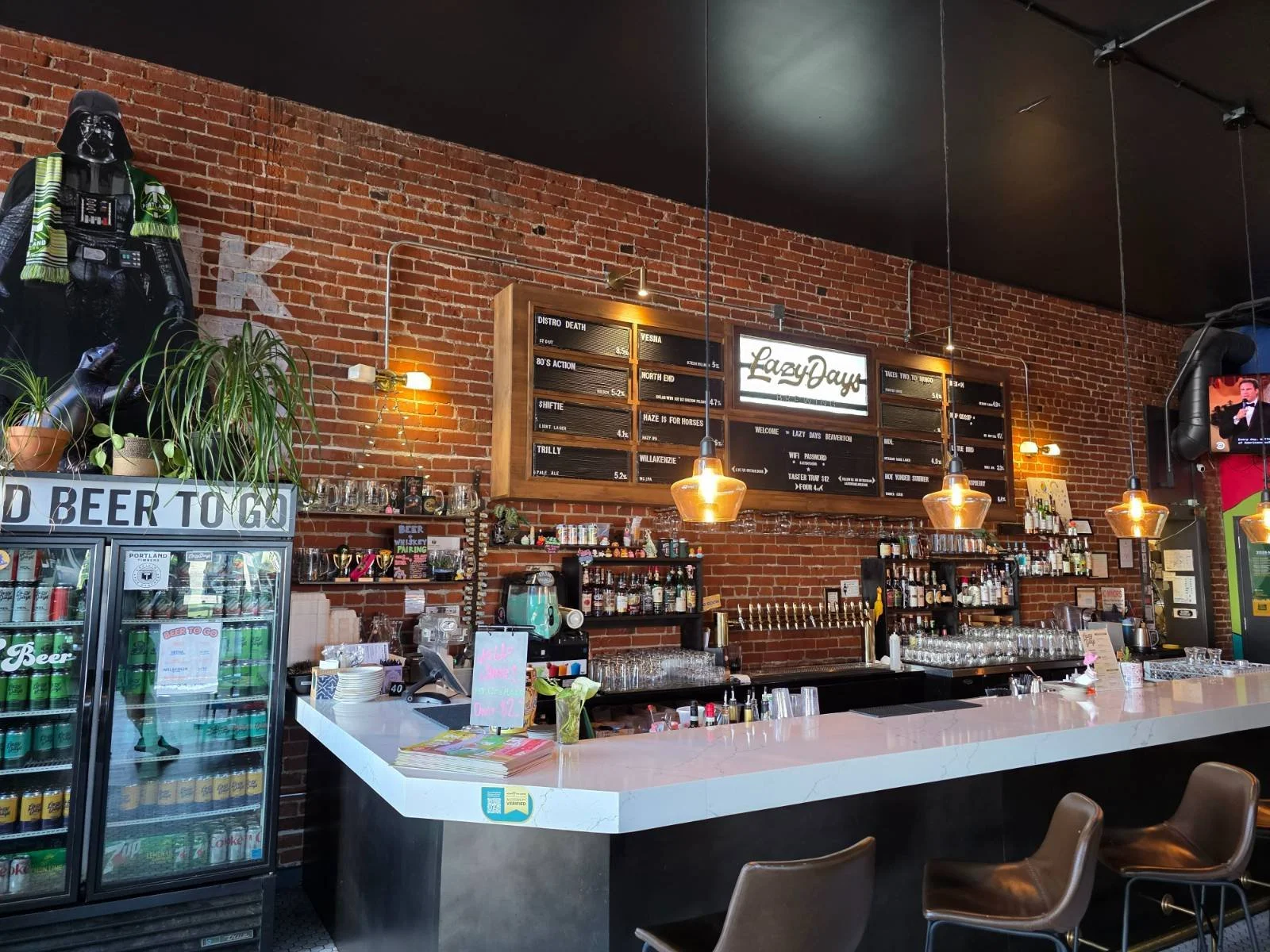 A bar in a craft beer brewery with several chairs at the bar, the wall behind the bar is brick and covered in beer menu and beer taps