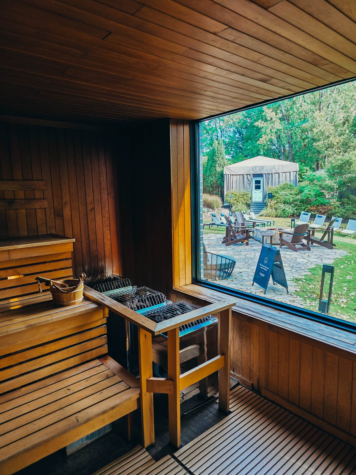 Inside a sauna with three tiers of seating and the stoves in the corner, to the right is a large glass window with a view of the lawn