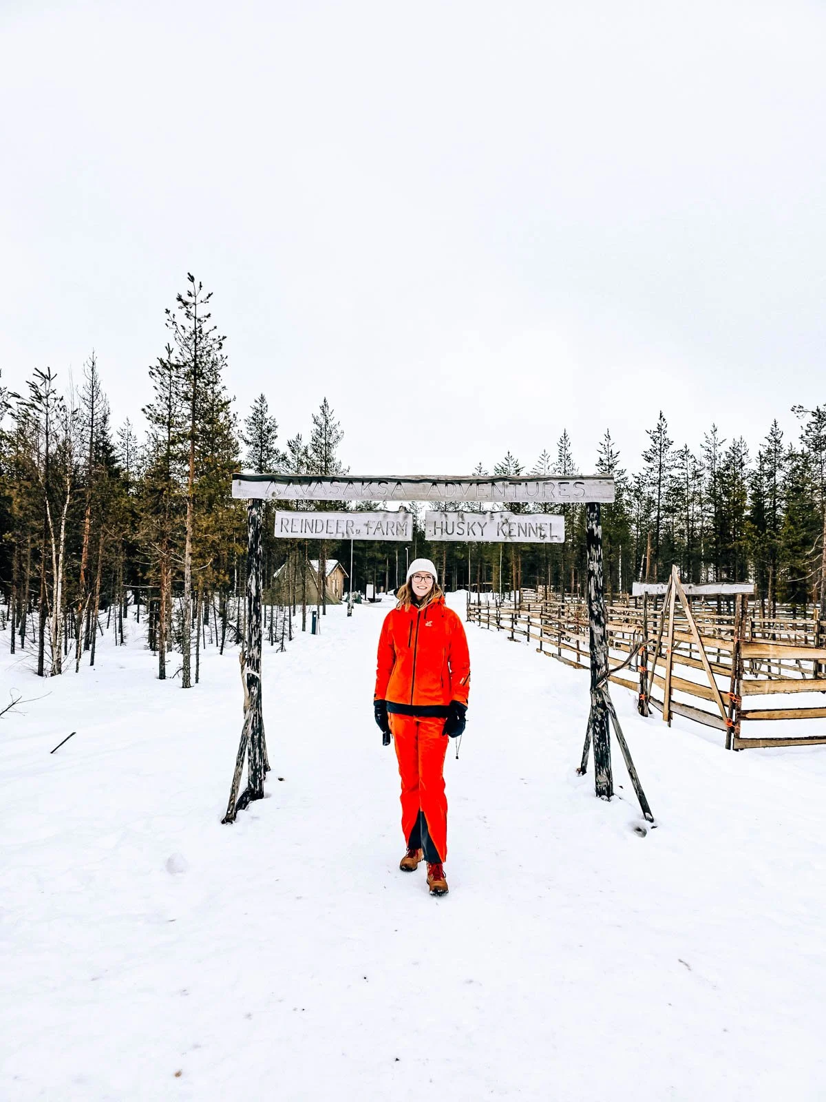 Helena in red standing under a wooden entrance sign for Aavasaksa Adventures in the snow with wooden fencing and green pine trees in the background