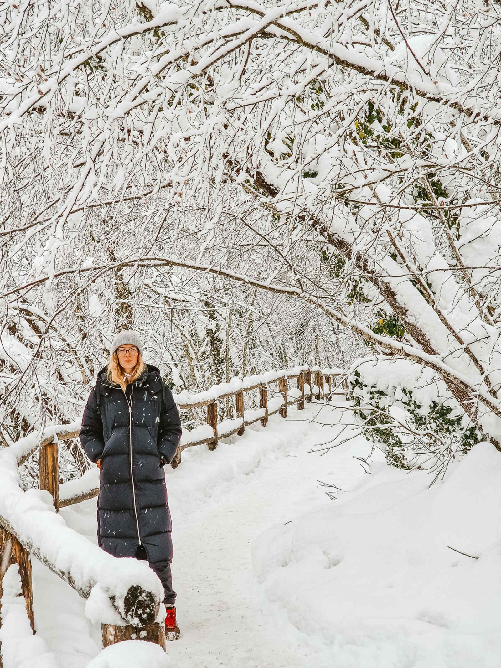 Girl in a long black puffy jacket standing on a snow covered path with snowy treeds and a snow covered fence next to the girl, snow in Plitvice Croatia