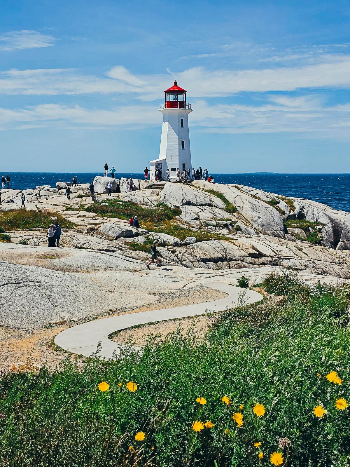a concrete path weaving over a rocky landscape towards a white lighthouse with a red roof. It's a clear, sunny day with blue sky and blue water behind the lighthouse where people are milling around on the rocks