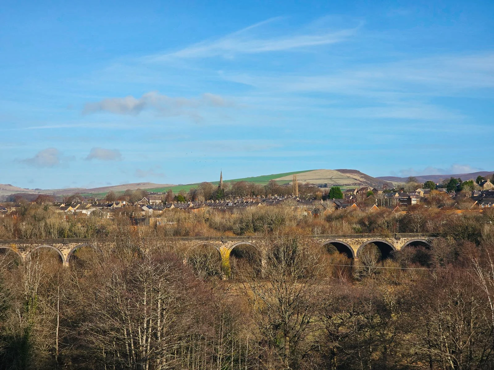 A panoramic view looking across a shallow valley with a stone viaduct in the middle, beyond it is a village with a church spire visible and green hills and blue sky beyond. A view along the Peak Forest Canal, an easy walk in the High Peak