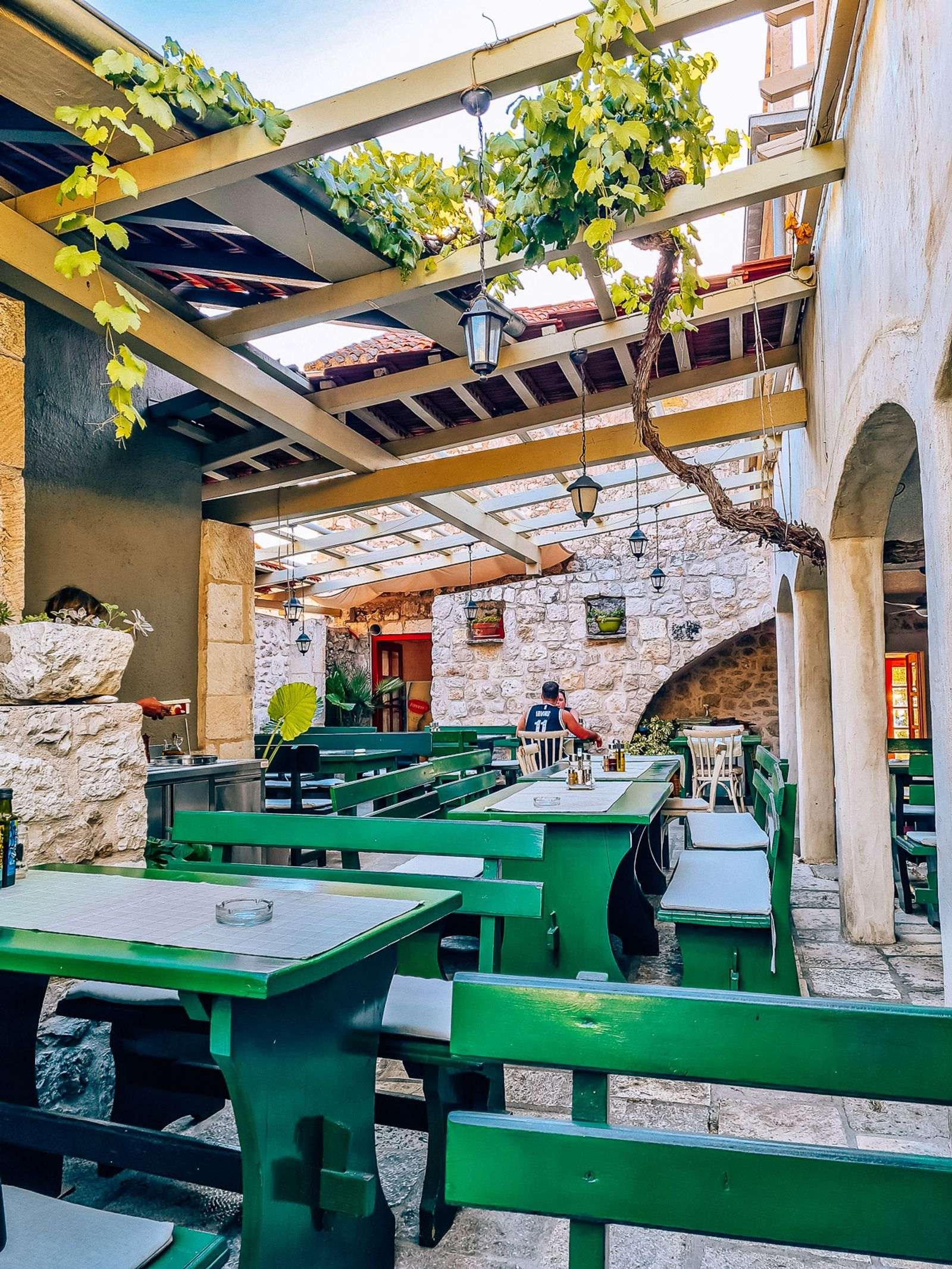 A quaint stone courtyard with a terrace and vines growing over the green wooden restaurant tables and benches