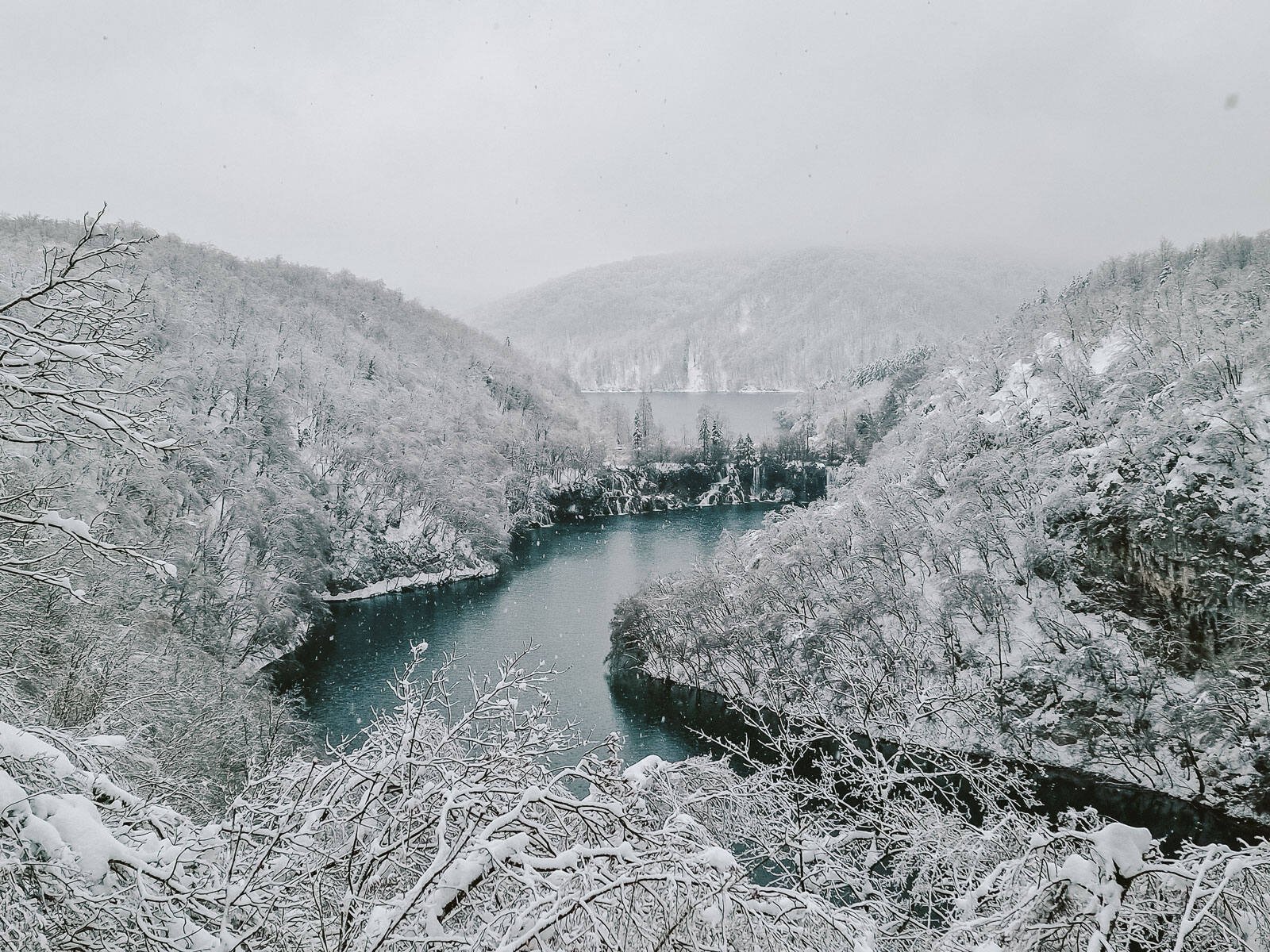 A panoramic view of Plitvice National Park in winter looking across a dark blue lake with snow covered trees on the hills each side, cloud is low hanging over more hills in the distance