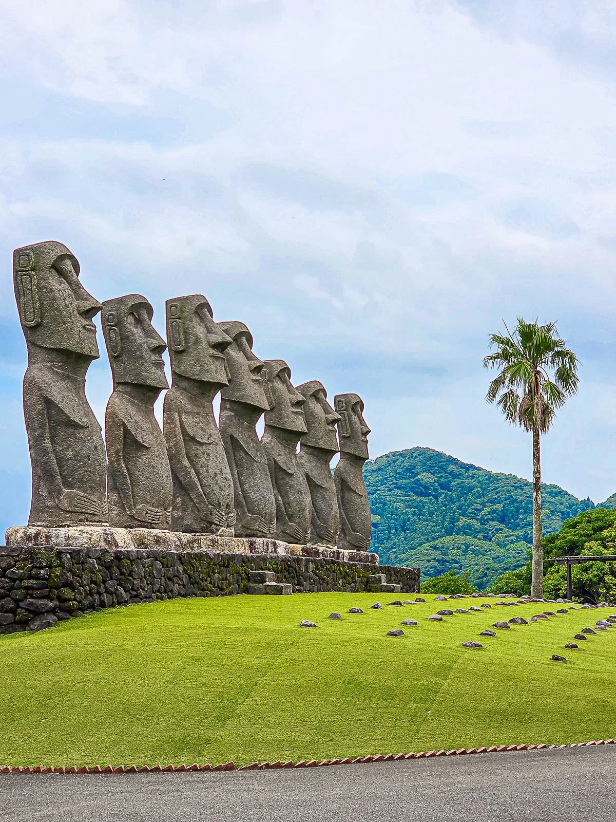 seven stone moai statues in a line with a neat, green lawn in front of them an a palm tree in the distance
