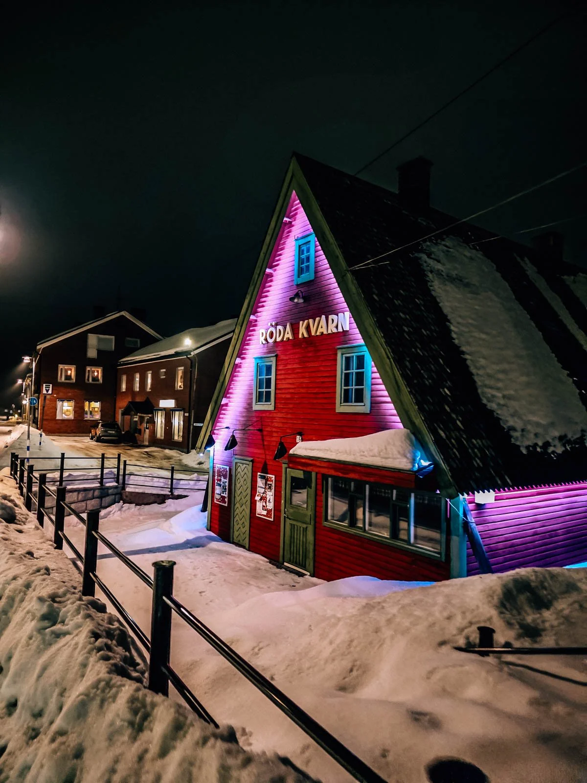 Red wooden building with green door and windows,  lit up at night in the snow in a town