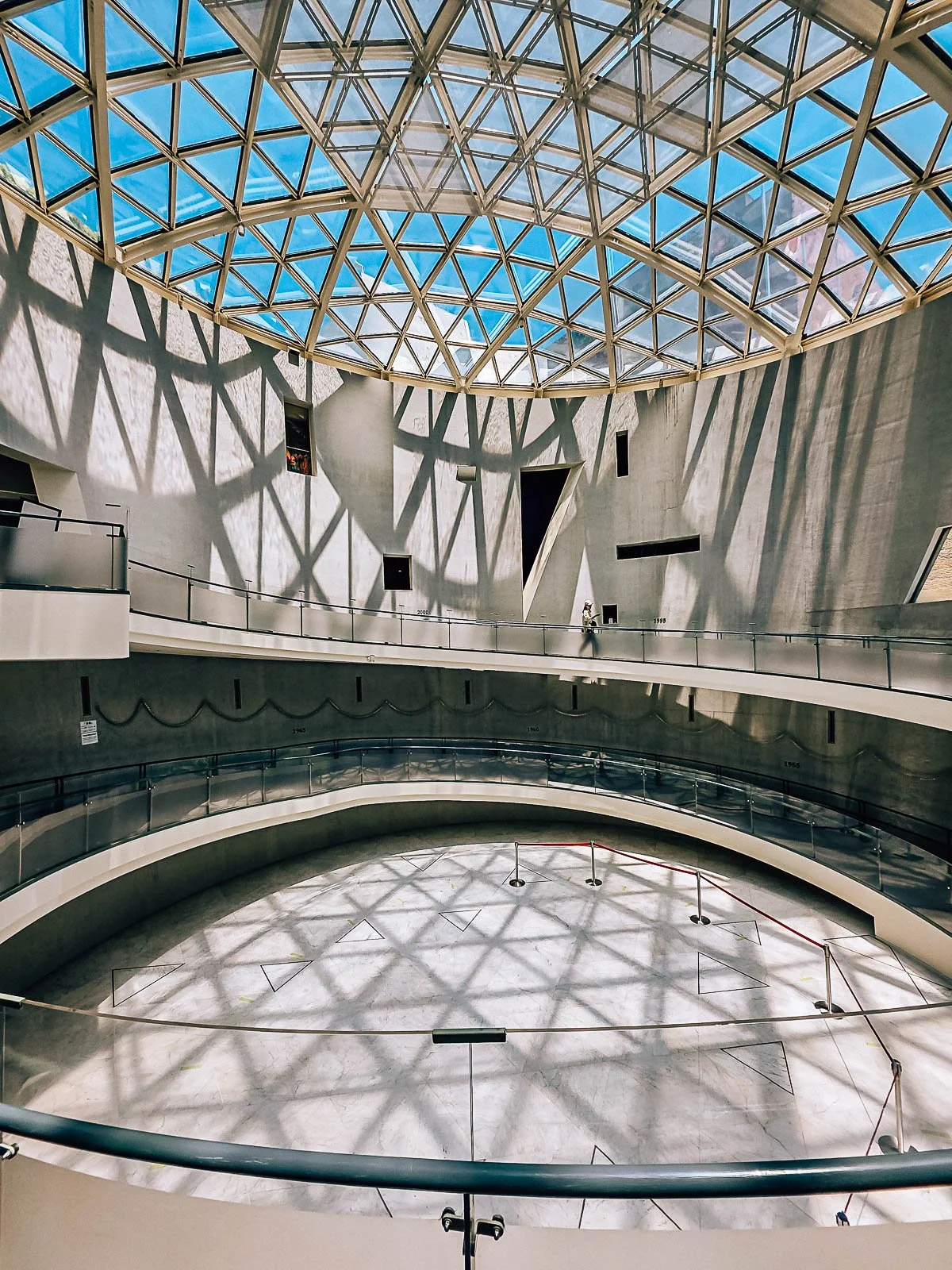 A large concrete circular atrium with a glass ceiling and sunshine coming through, showing the triangluar pattern of the glass roof on the floor. A narrow walkway runs in a circle along the edge of the wall with one personal walking along it