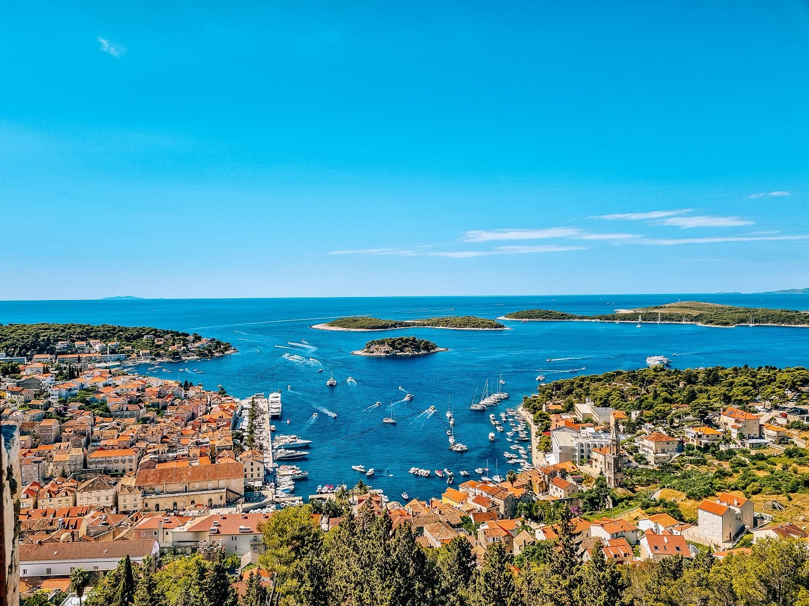 view from the fortress of Hvar Town below with the harbour and many boats sailing in and out. The sea is blue and there are green islands in the distance