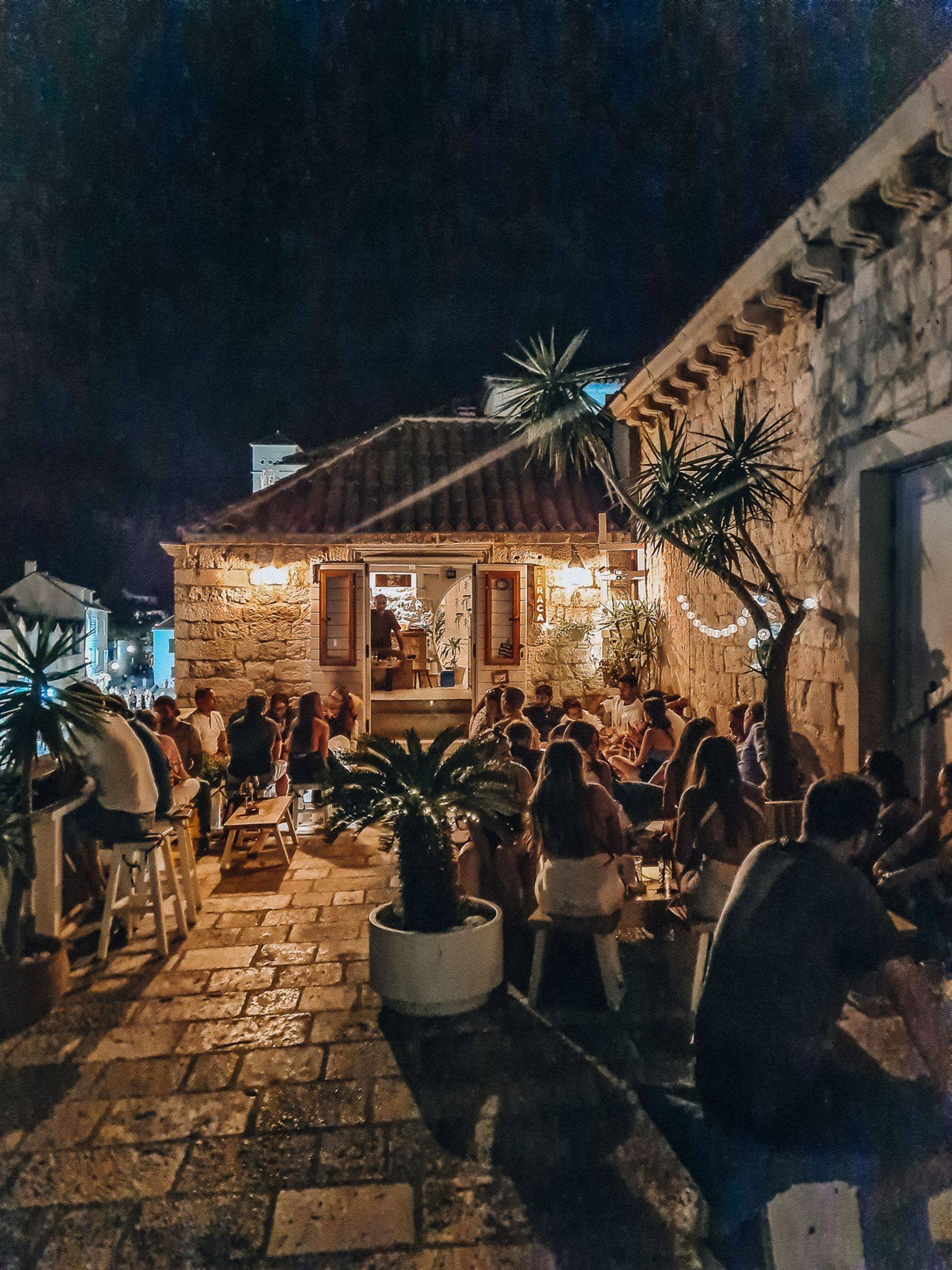 A night time shot of a stone building which is a bar and terrace with many people drinking in the low light