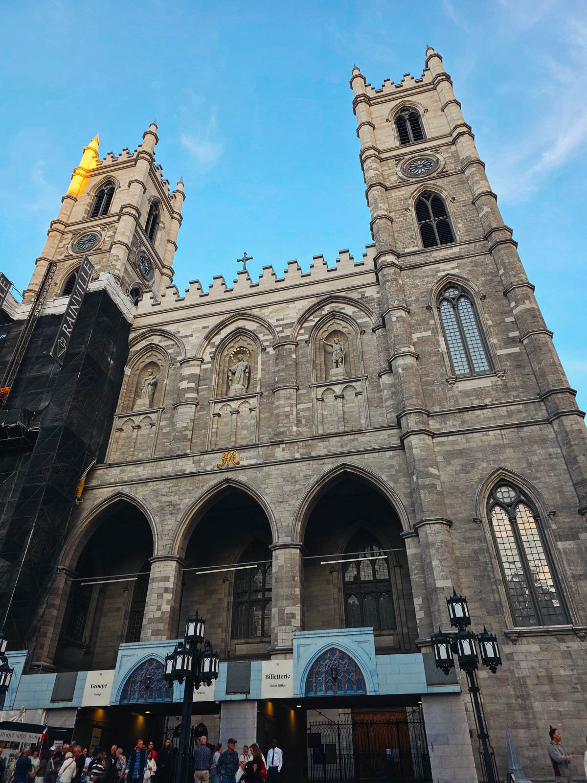 The Notre Dame Basilica Cathedral in Montreal, made of stone with two towers on the front and a clock face on each. There are many ornate arches and arched windows
