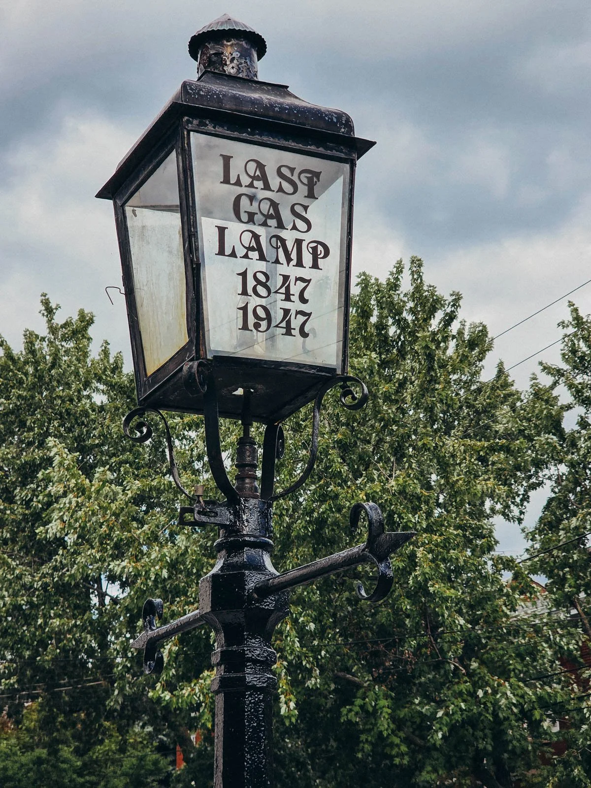 A large old black gas lamp outside with trees in the background, the gas lap has writing on that says "last gas lamp 1847-1947" in Kingston Ontario