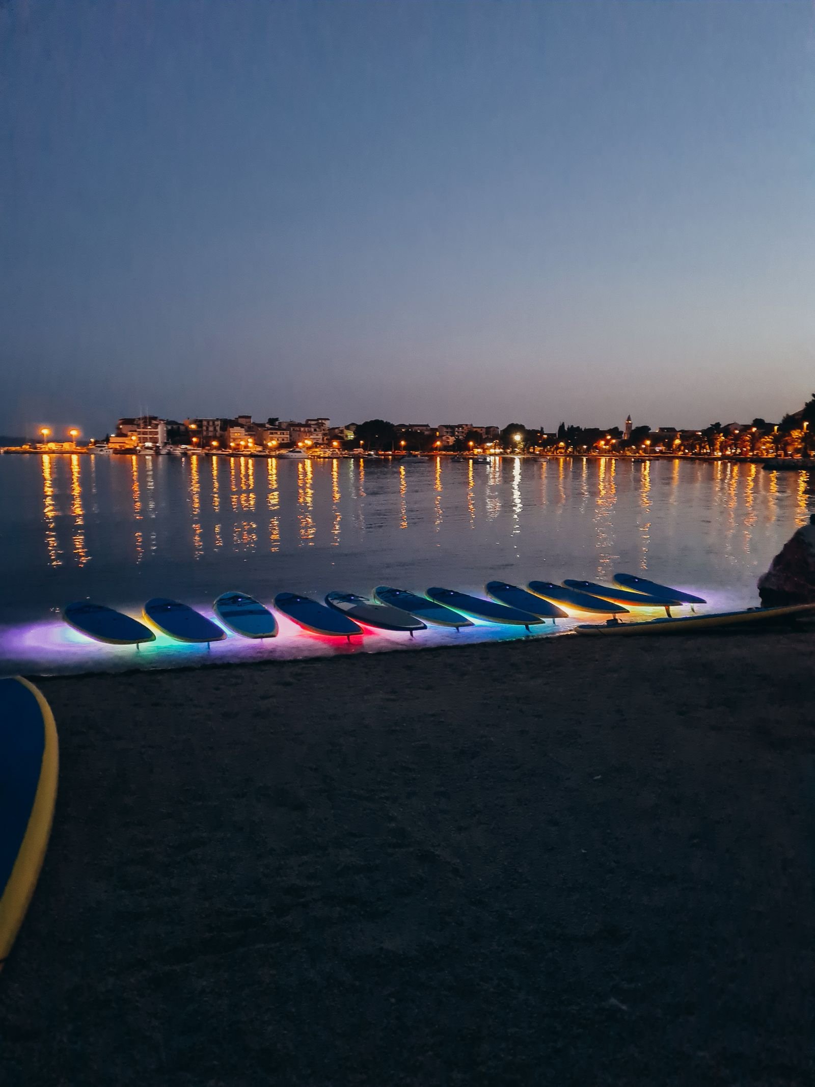 paddleboards lined up on the beach at dusk with neon lights underneath them