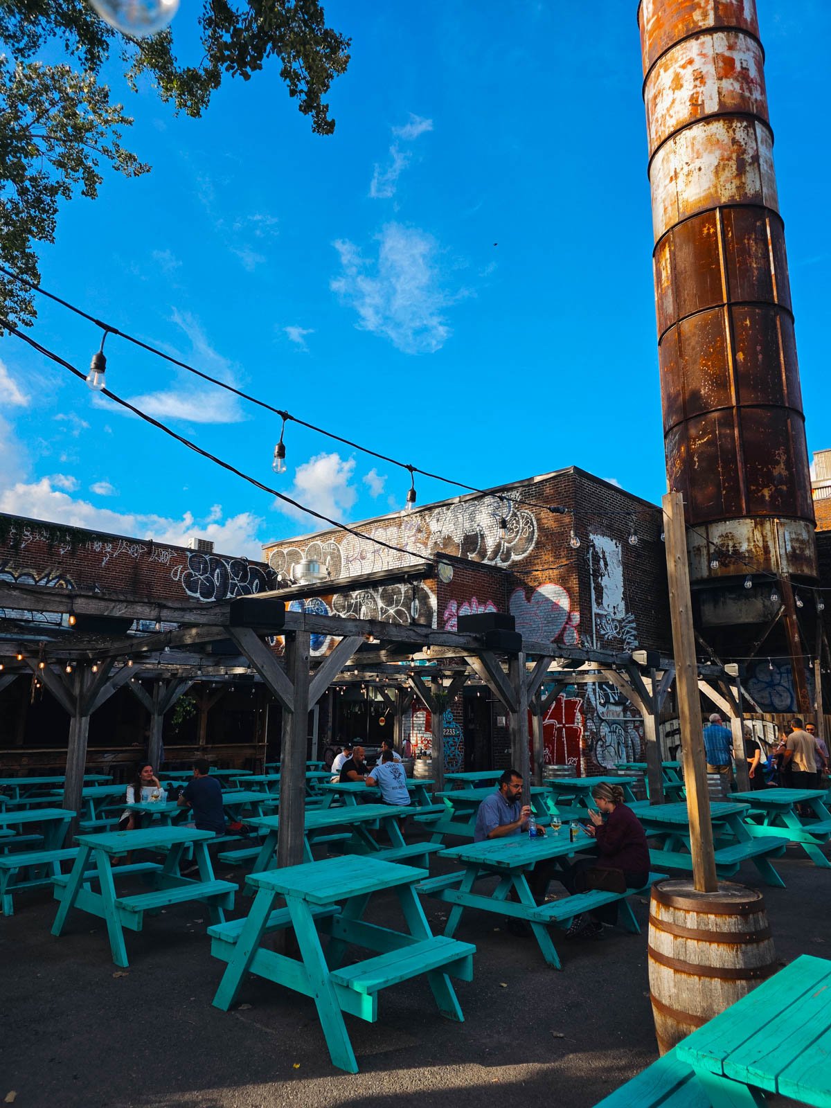 An urban industrial area with lots of graffiti on buildings and string lights and pergolas with people sitting at the many turquoise picnic benches underneath. This is Messorem brewery in montreal