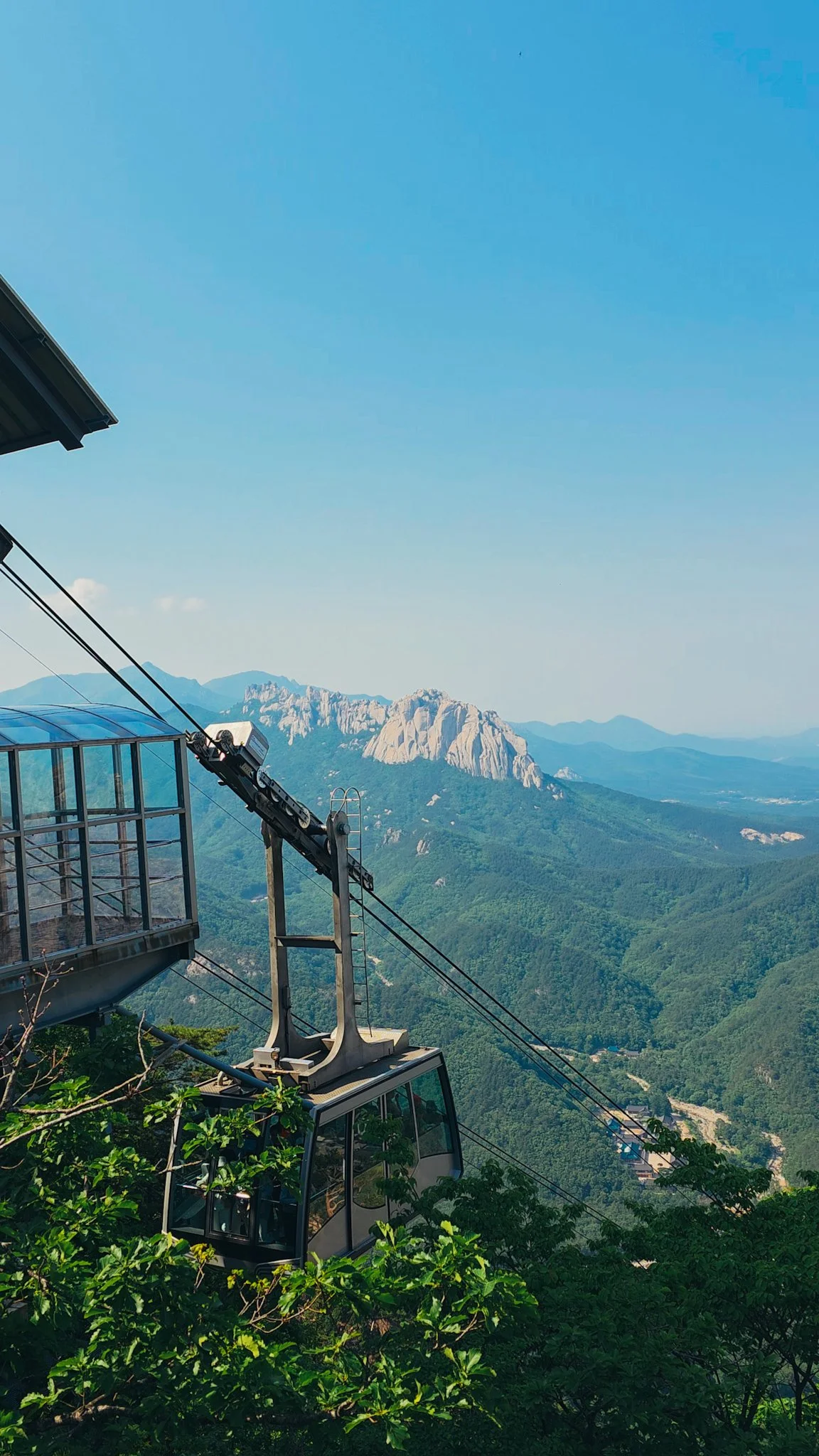 A large mountain cable car just leaving the cable car station to descend the mountain, surrounded by green trees. In the distance is a rocky mountain peak