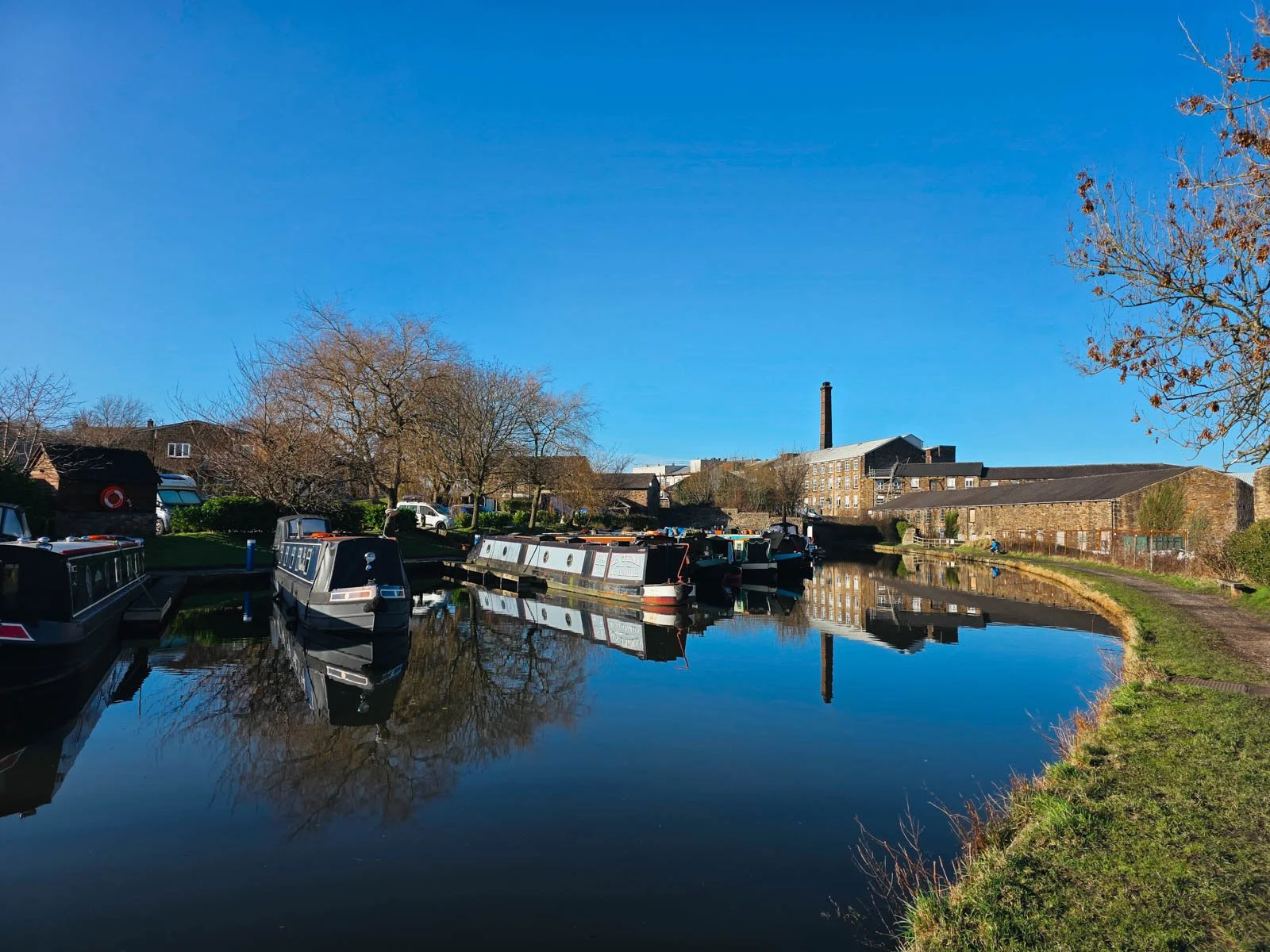 A canal marina with many canal boats lined along the left, on the right is the canal path leading to old stone buildings. It's a clear sunny day