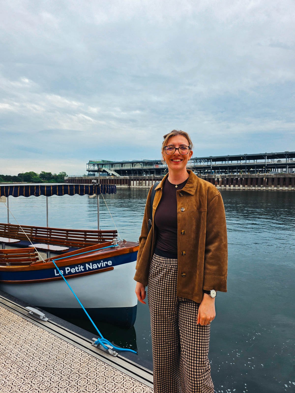 Helena in a brown jacket and checked trousers standing in front of a small wooden boat that says "Le Petit Navire" on the side