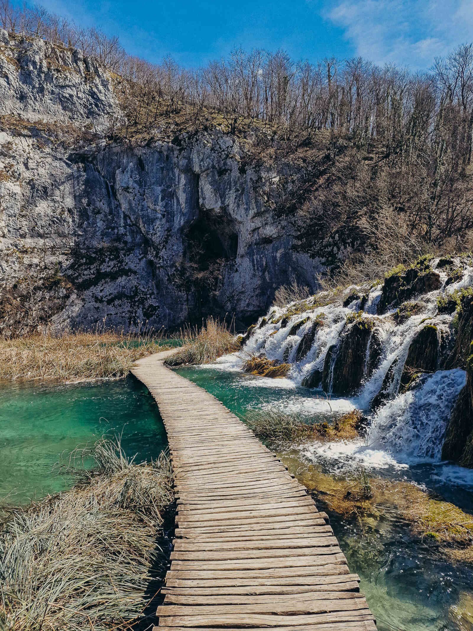 a wooden walkway over turquoise water with small waterfalls running on the righthand side. Cliffs with a cave are in the distance