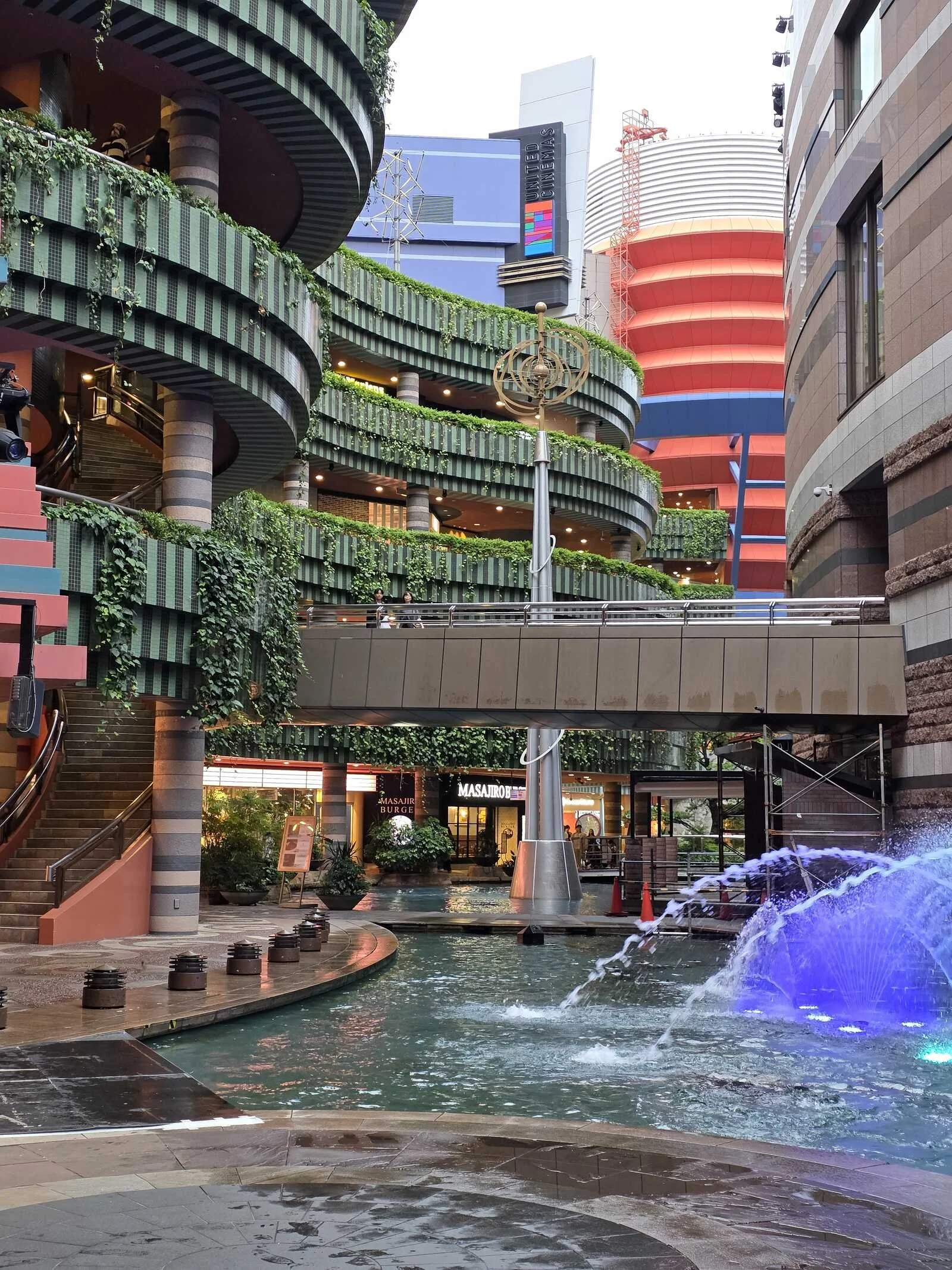 An open air mall with greenery over green balconies on multiple levels and a fountain in the middle
