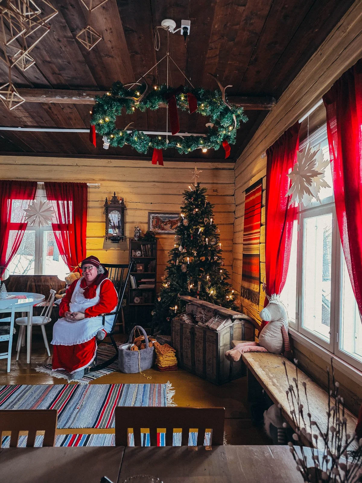 A Mrs. Santa in red sitting in a rocking chair inside a wooden cabin decorated for Christmas with a large Christmas tree and red curtains