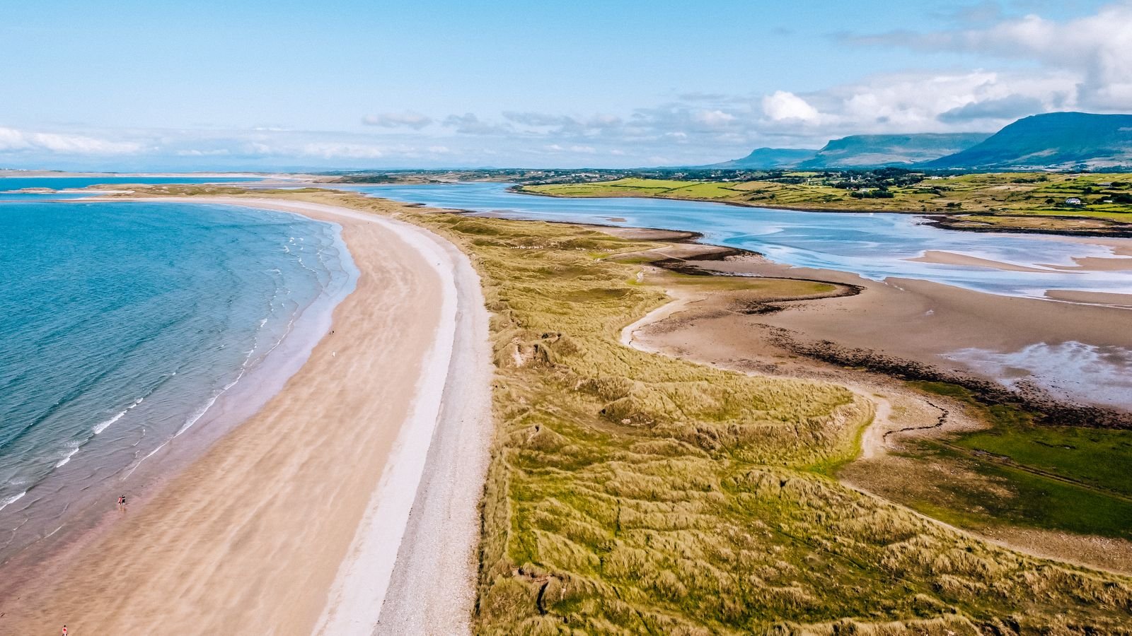 drone photo above a long stretch of beach with blue water and grassy sand dunes with mountains in the background