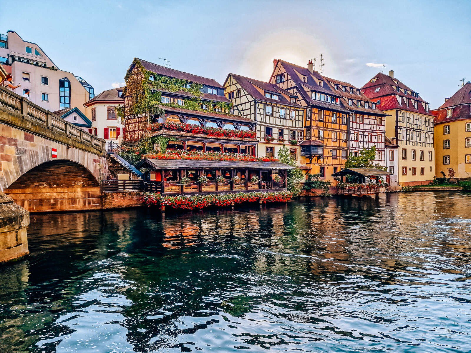 A river view with one side of the bank lined with many colourful old timber buildings. One of the buildings lined with many flowers and plants