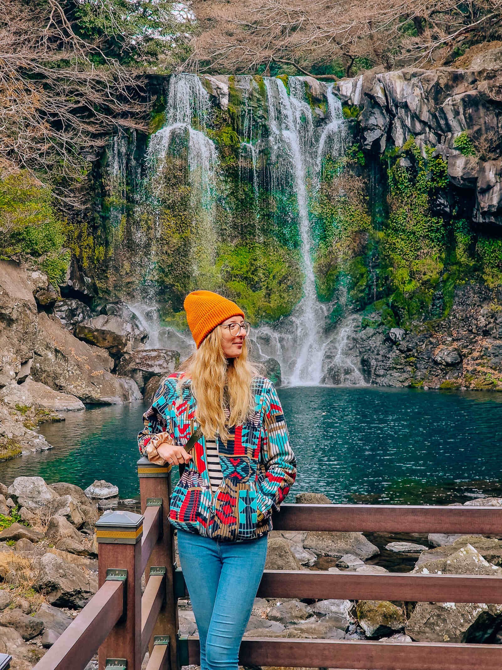 woman in a brightly patterned fleece and orange hat standing on a wooden deck with a waterfall flowing down a green moss covered cliff into a dark blue pool