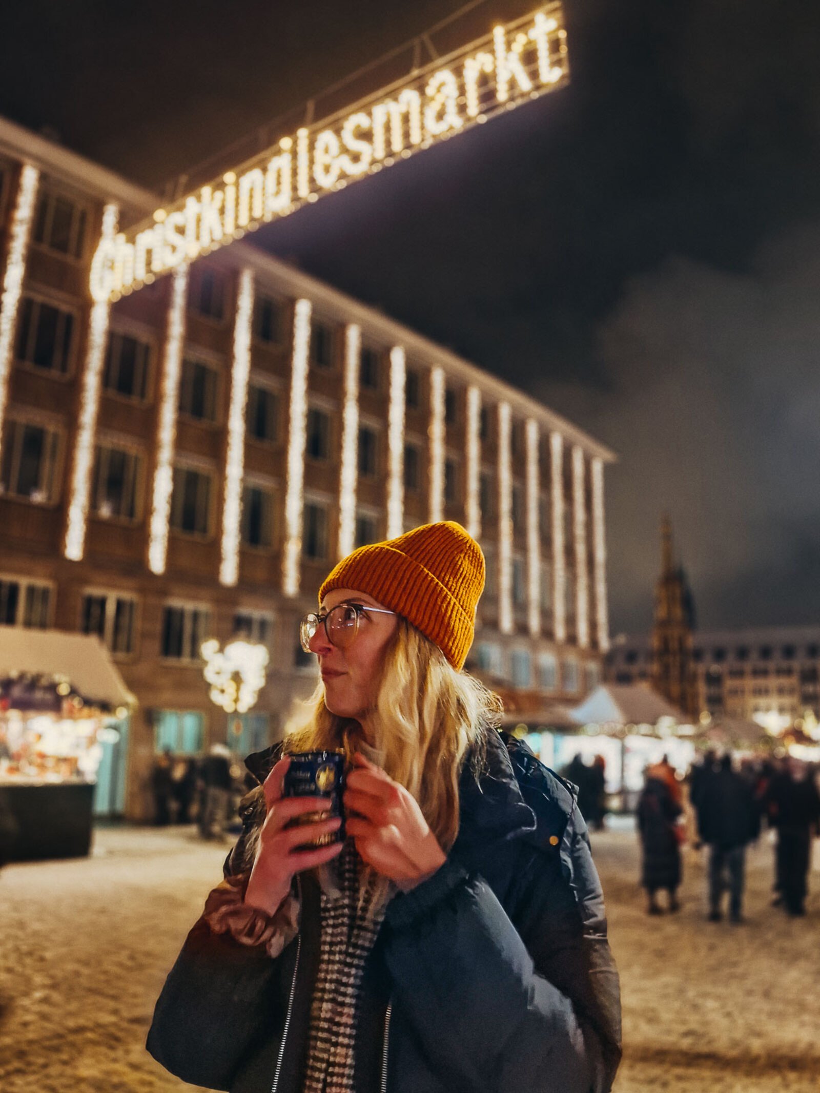 girl holding a mug inder an illuminated sign that says Christkindlesmarkt