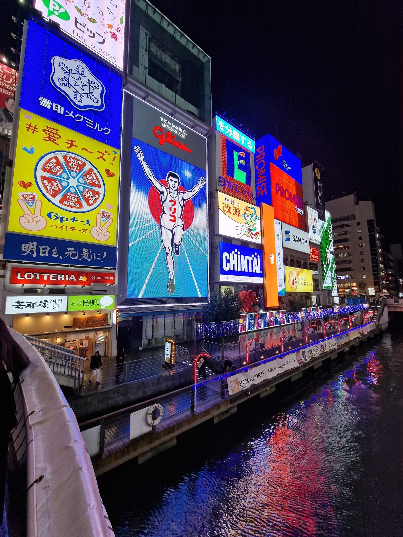 Nighttime with Lots of colourful adverts and billboards lit up on the side of buildings along a river including a large portrait of a running man with his arms in the air - this is the osaka glico running man sign