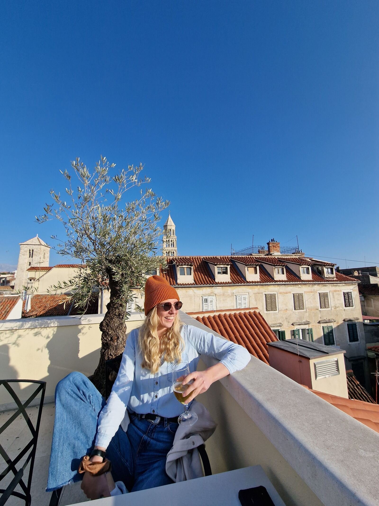 Woman in a blue shirt and denim jeans, holding a beer and leaning on a low wall on a rooftop with views of old houses and terracotta roofs around her
