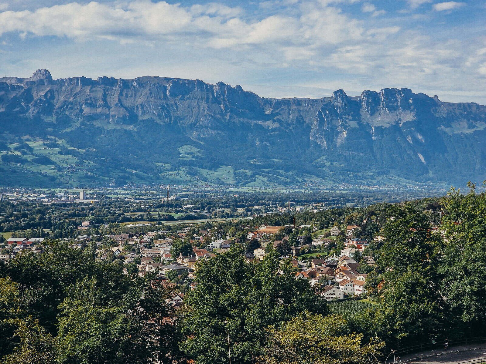 A panoramic view of Liechtenstein with lots of greenery, a town in the distance and mountains in the background
