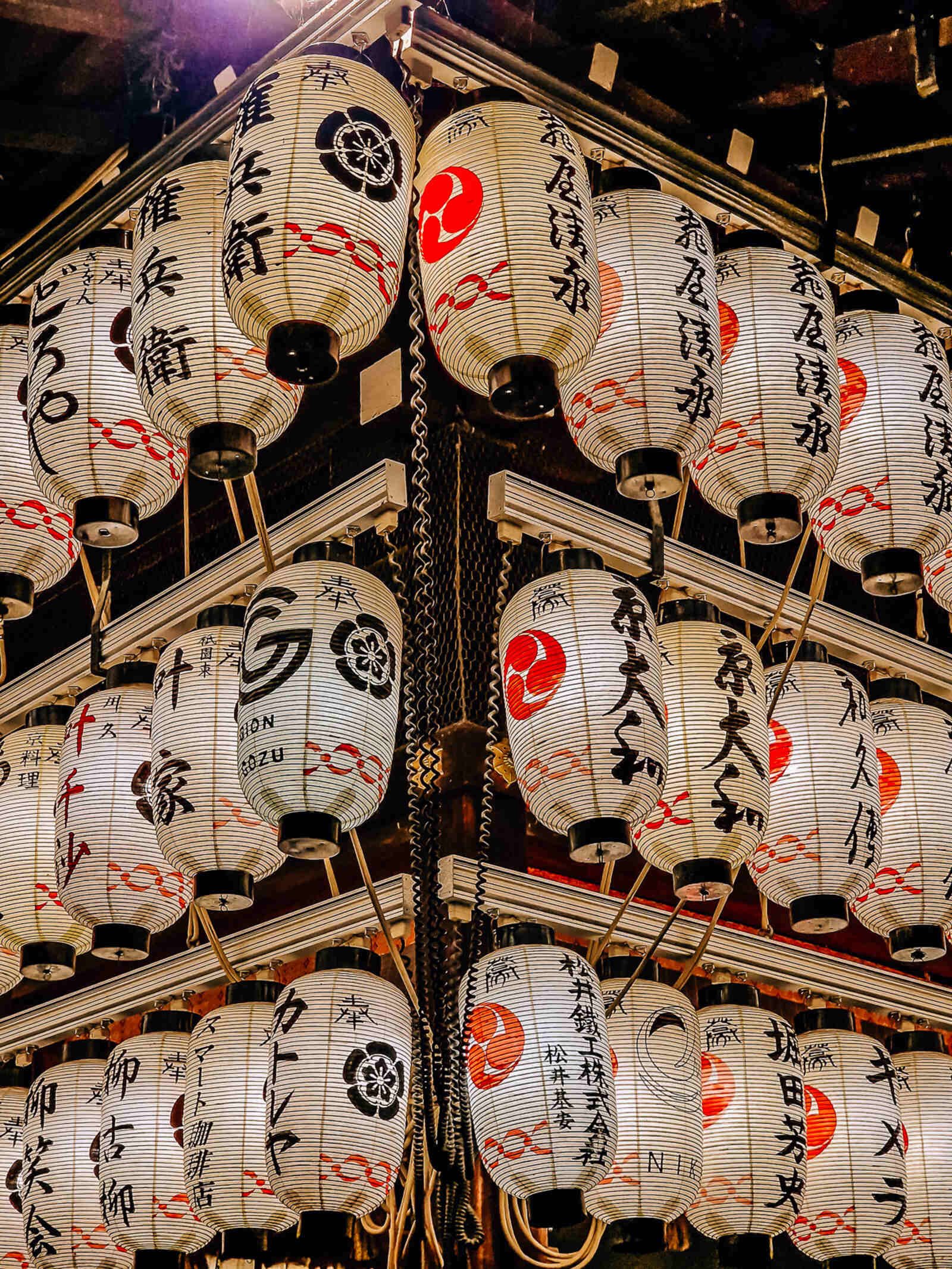 a wall of white Japanese lanterns lit up at night with black and red Japanese writing on them