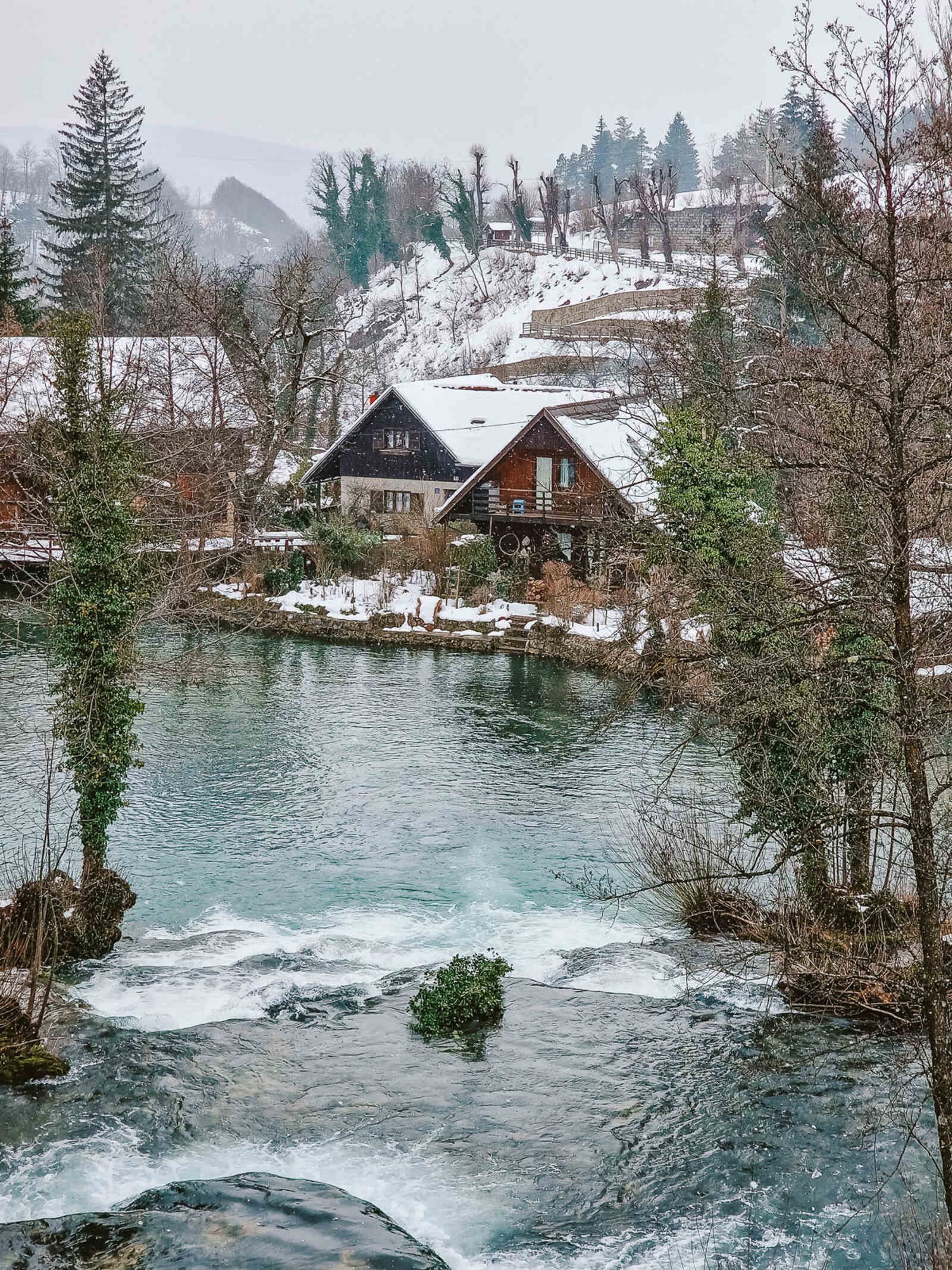 wooden a-frame cabins on the river banks in rastoke village, roofs are white with snow and snow is falling. Hills behind the houses are covered in snow too