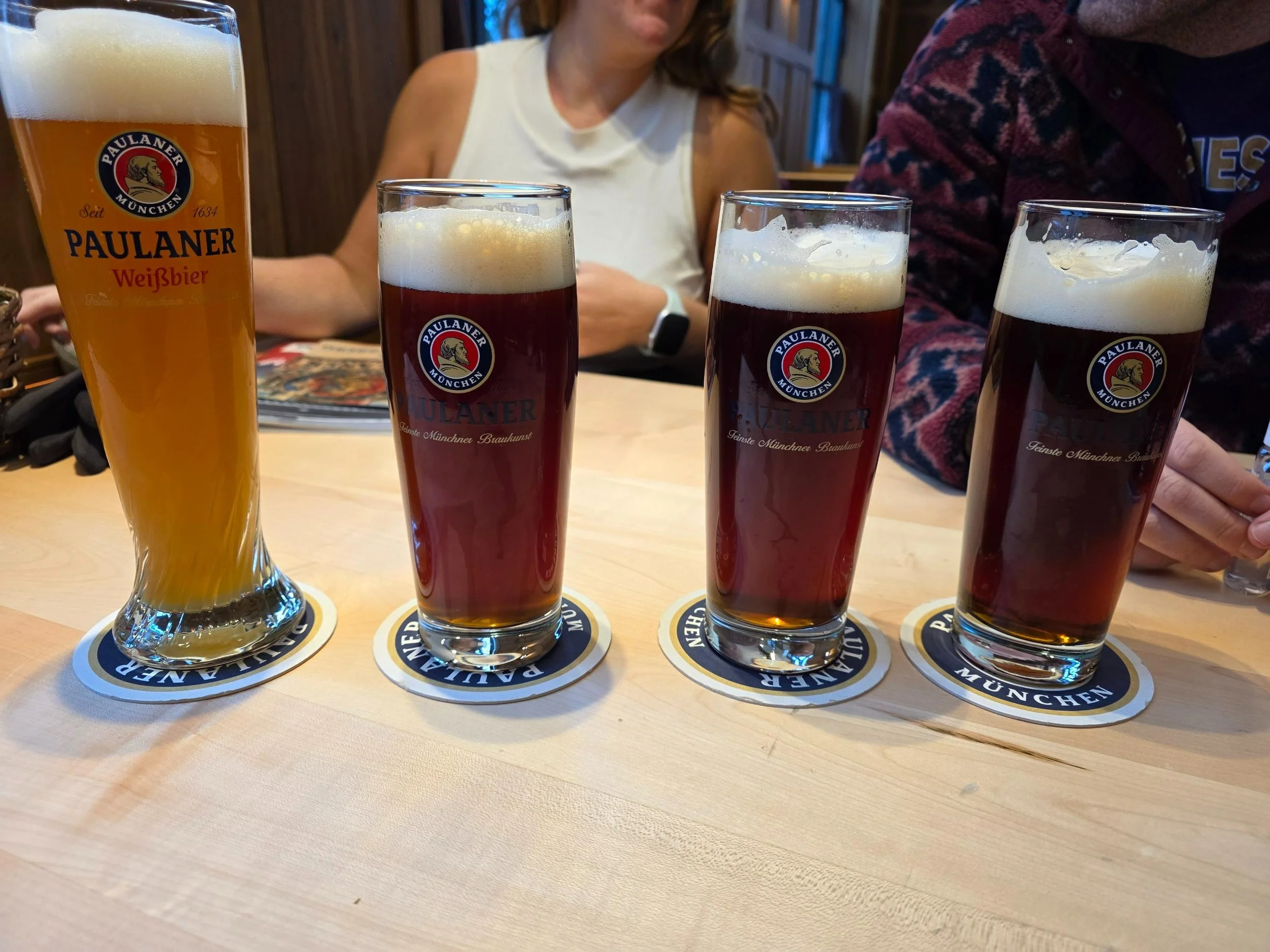 Two people sitting at a table with for large beers infront of them at a beer hall. Three of the beers are a dark amber colour and one is a golden colour