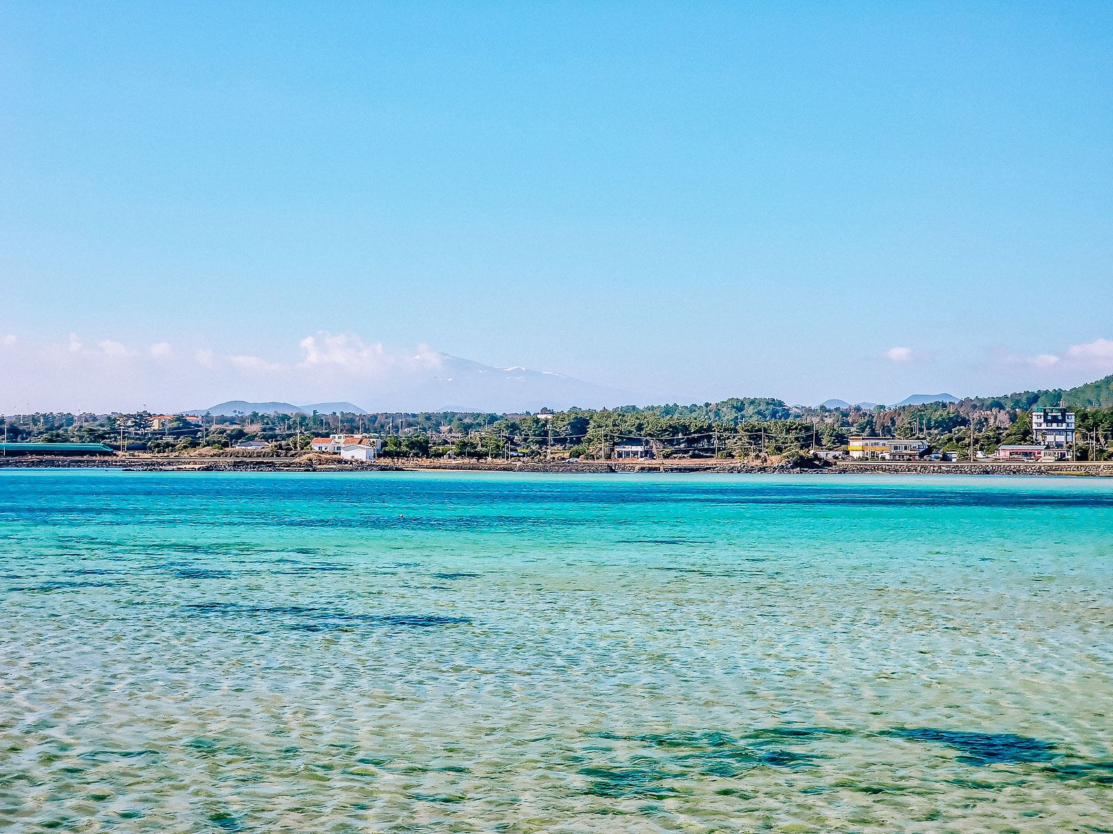 calm, crystal clear turquoise waters in a bay with a small town and trees visible on the other side