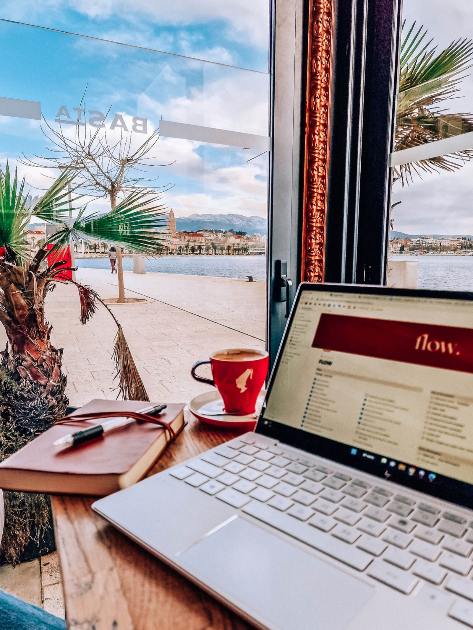 A laptop open on a cafe table with a red coffee cup and leather notebook next to it, the view out the window behind is of a coastal city harbour, Split Croatia
