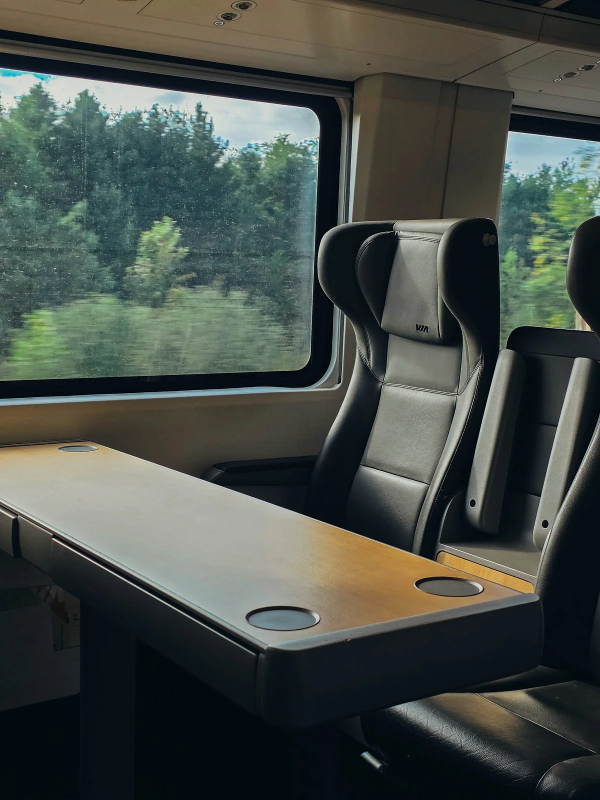 A train table and seat in business class on VIA  Rail with dark grey leather chairs and a wide table with cup holders. A lush green landscape is out the window