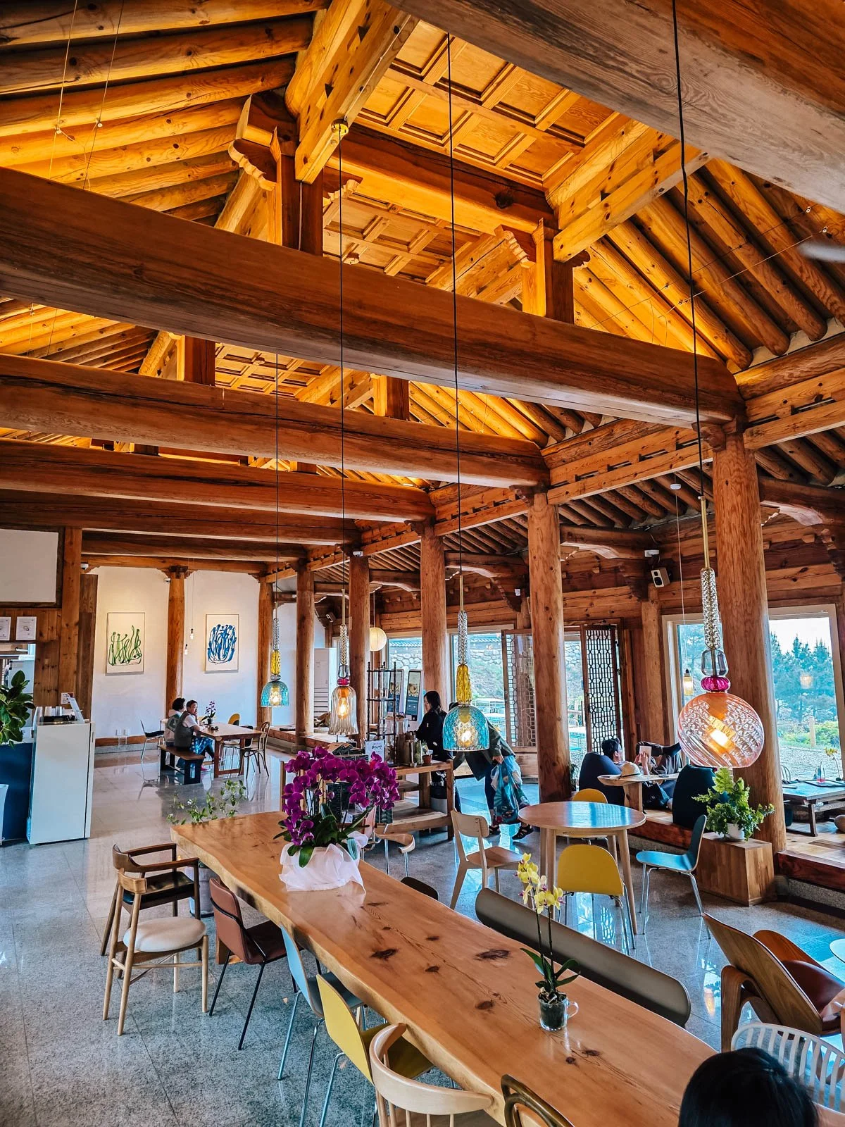 Inside a cafe made of warm wood with a vaulted ceiling and large windows on the walls. There are many tables in the room, some of them occupied with people and colourful glass lamps hanging from the tall ceiling